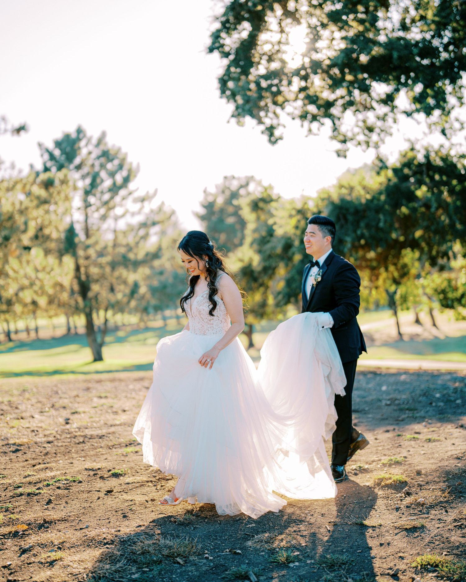 A couple walks through sunlit trees in wedding attire with a flowing white dress.
