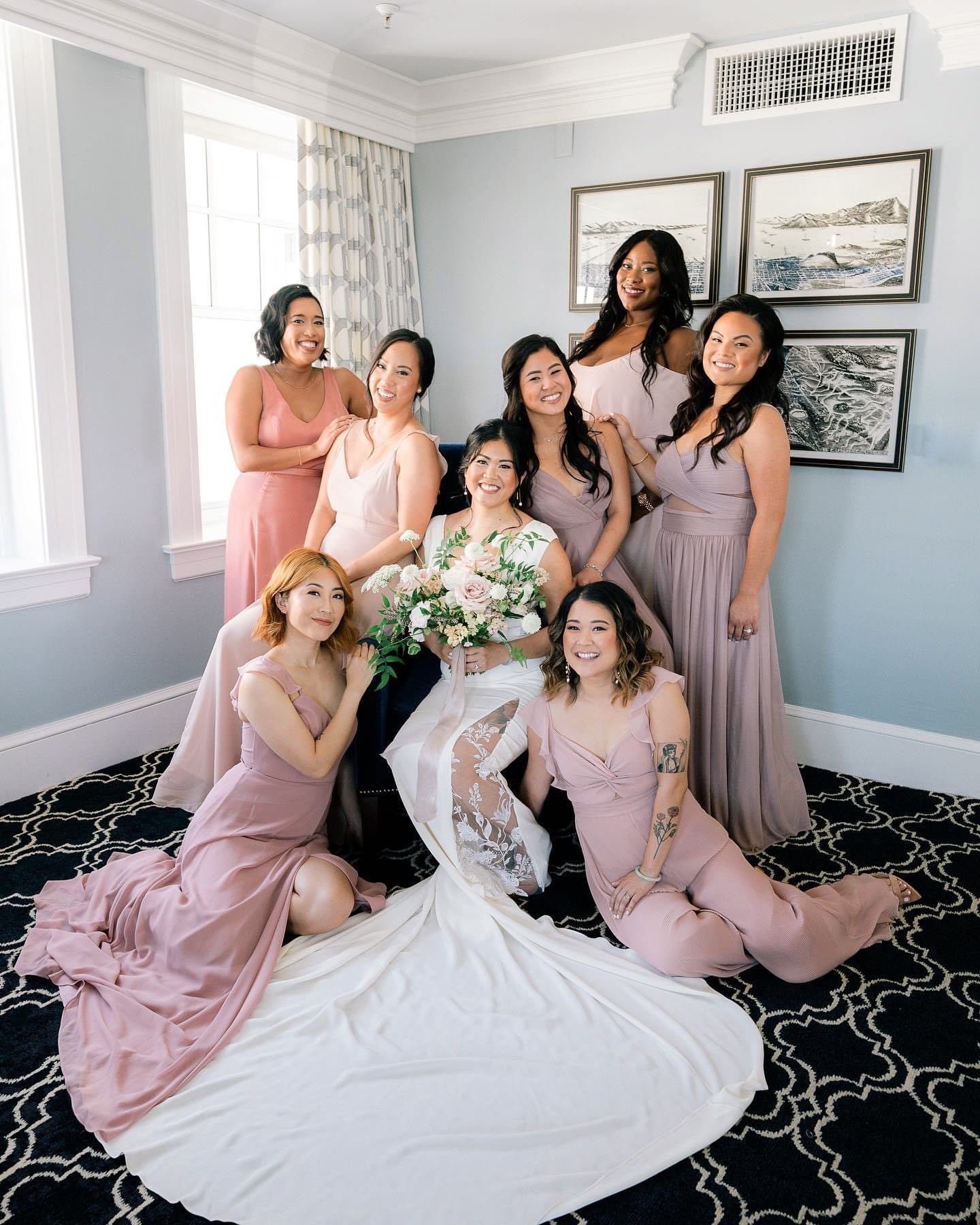 A bridal party in blush pink dresses poses together in a hotel room with decorative artwork.