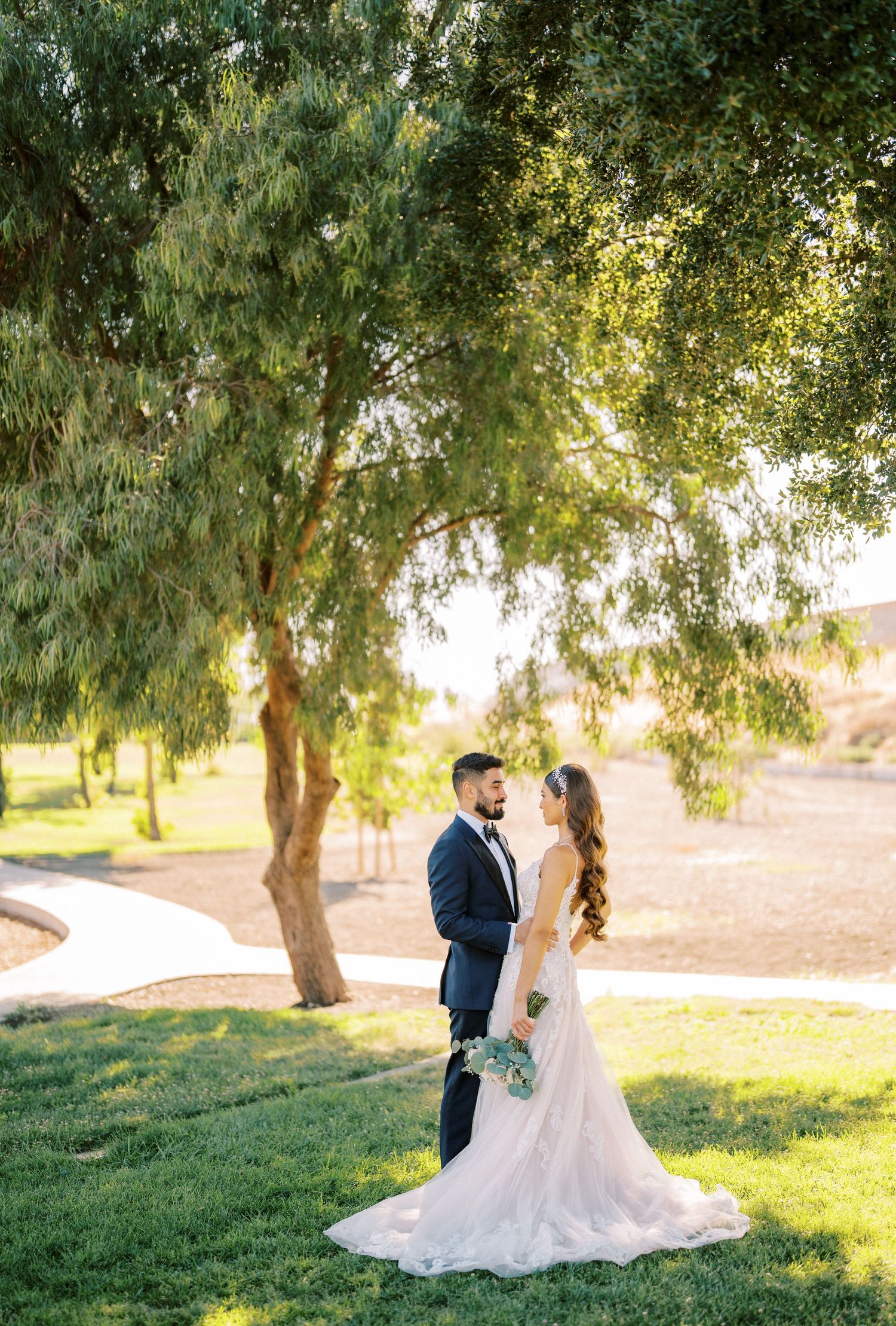 A couple in wedding attire embraces under a willow tree in a sunlit garden setting.