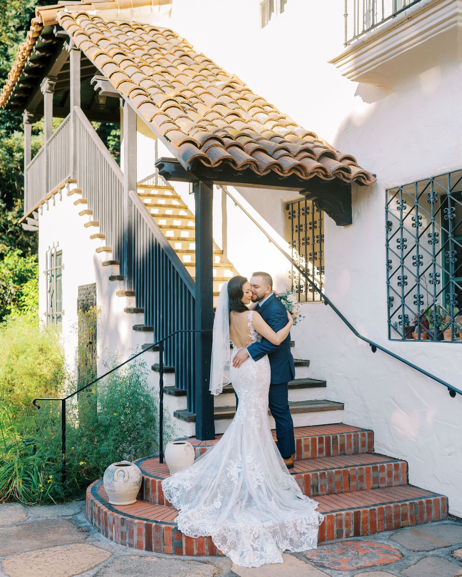 A wedding couple poses on Spanish-style steps with a long flowing white dress train.
