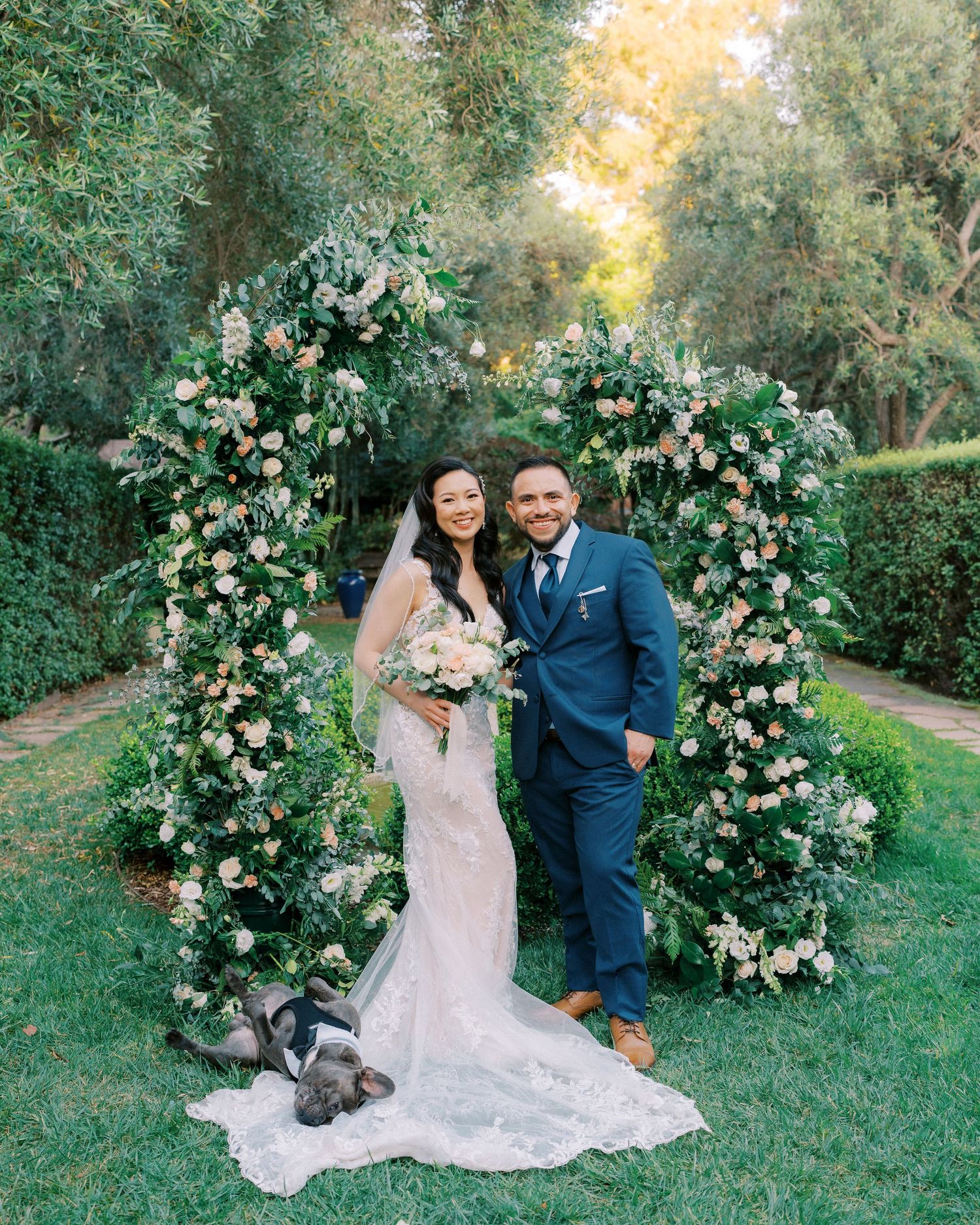 A wedding couple stands between two floral arches filled with white roses and greenery in an outdoor garden setting.