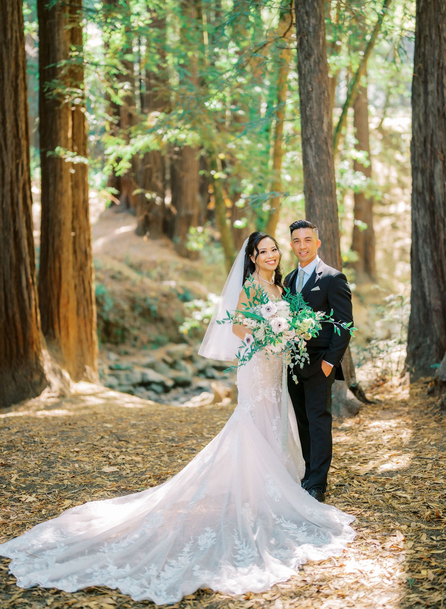 A couple poses in wedding attire among towering redwood trees in a forest.
