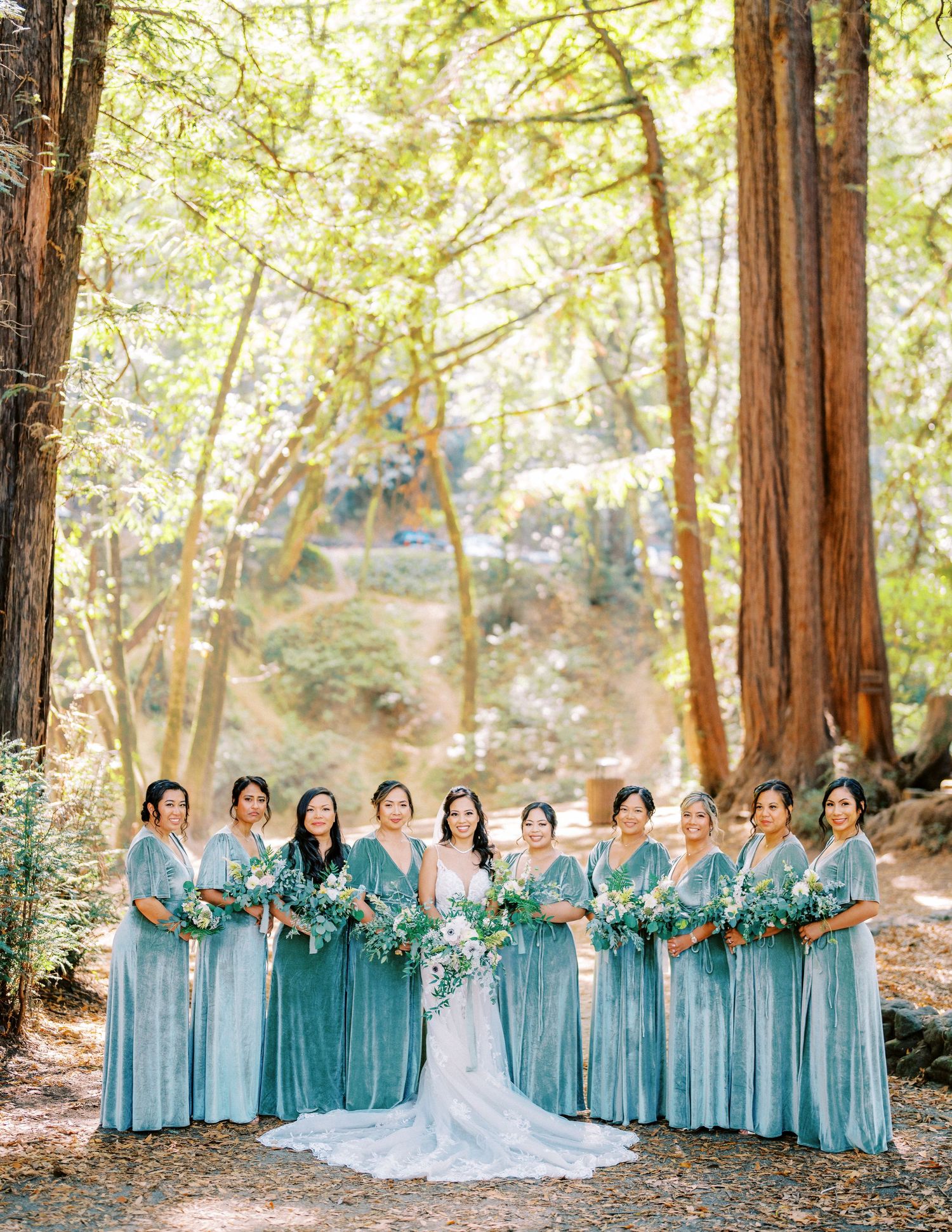 A bridal party in turquoise dresses poses together in a sun-dappled forest setting.