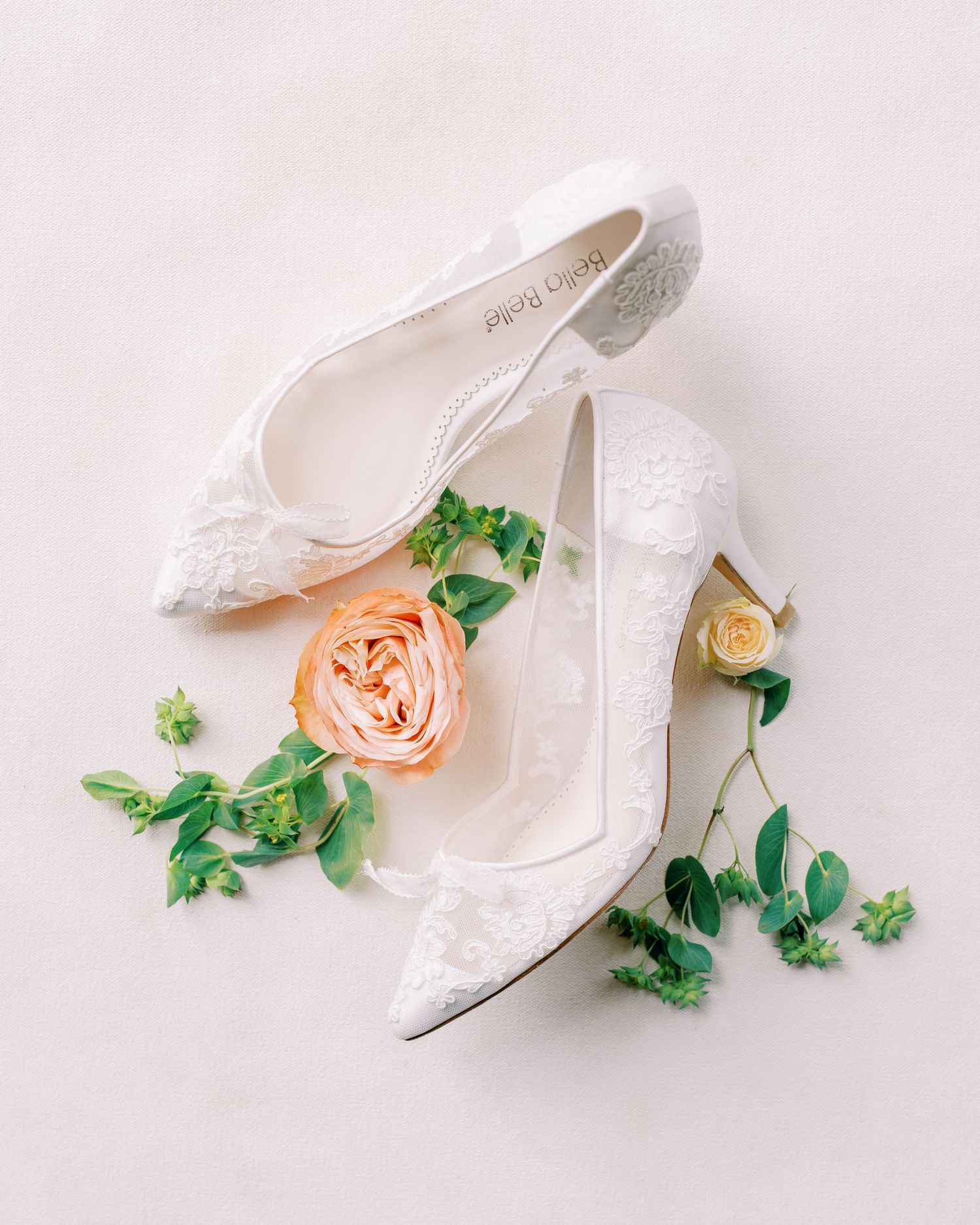 Elegant white lace wedding shoes with peach roses and greenery on light pink background.