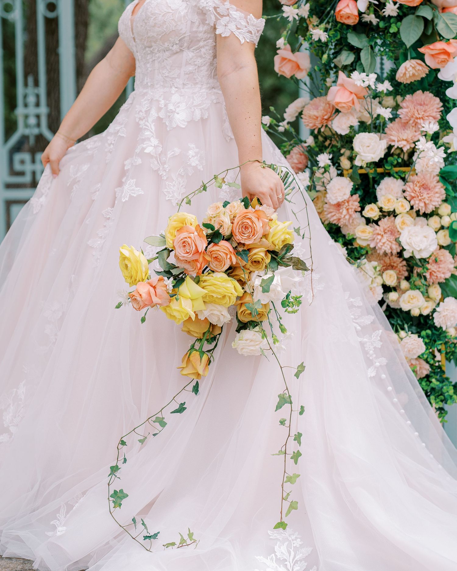 A flowing blush wedding gown with delicate lace details and cascading peach and yellow bouquet.