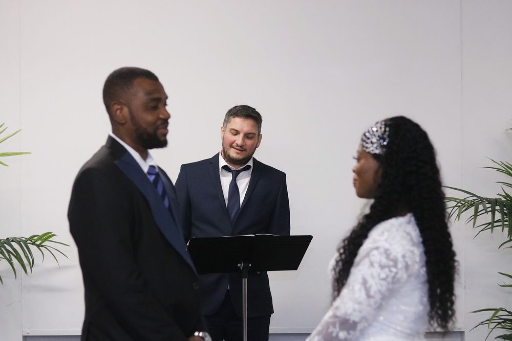 Wedding ceremony with couple and officiant standing between palm plants against white backdrop.