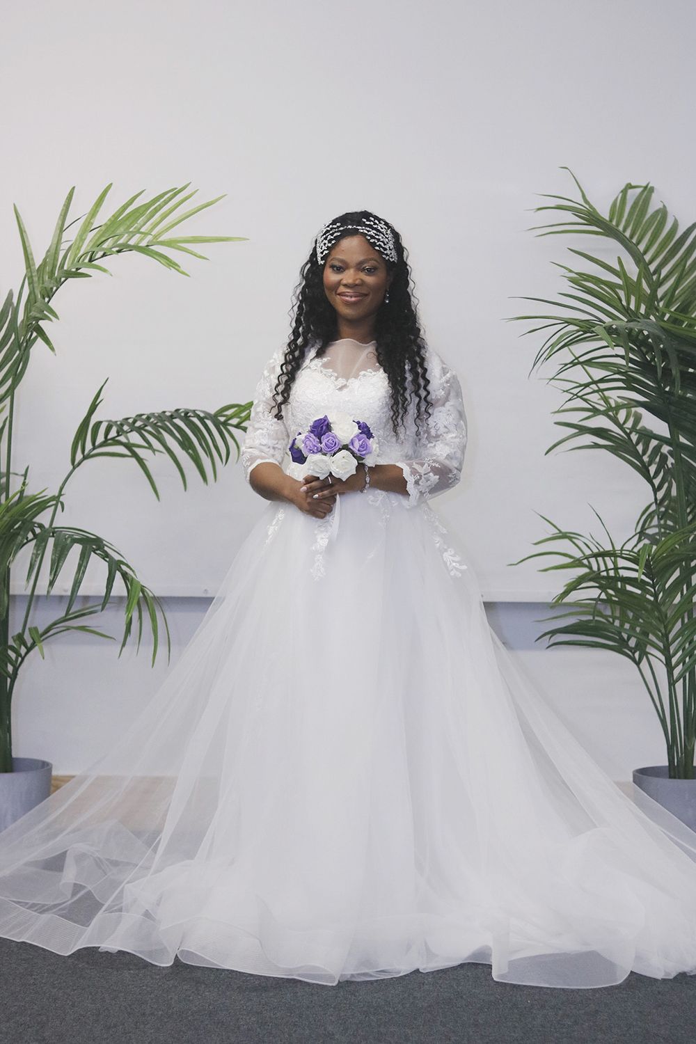 Bride in flowing white gown and crystal headpiece holding purple bouquet between tropical plants.