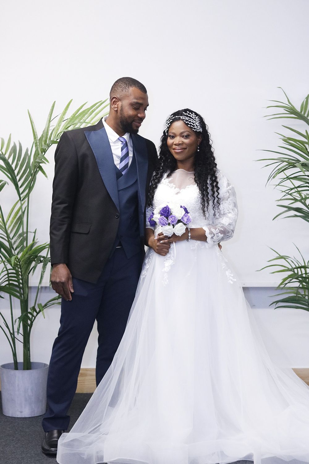 Wedding portrait with couple in navy suit and white gown holding purple flowers against palm backdrop.