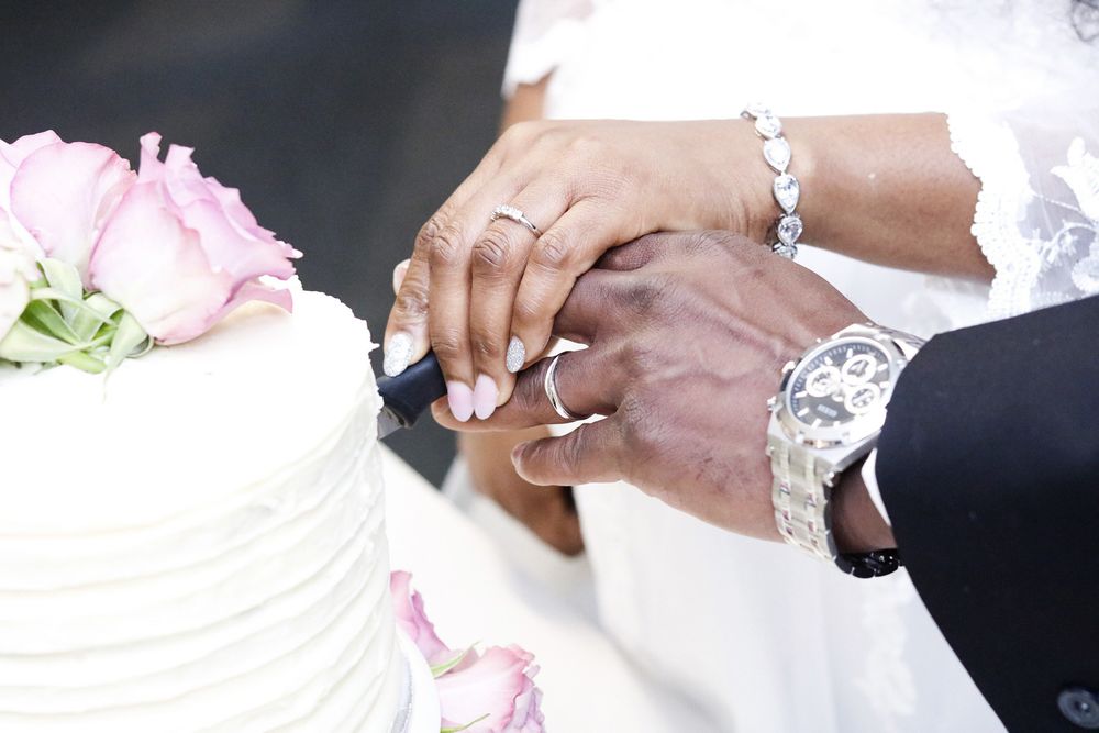 Close-up of hands cutting a white wedding cake decorated with pink roses.
