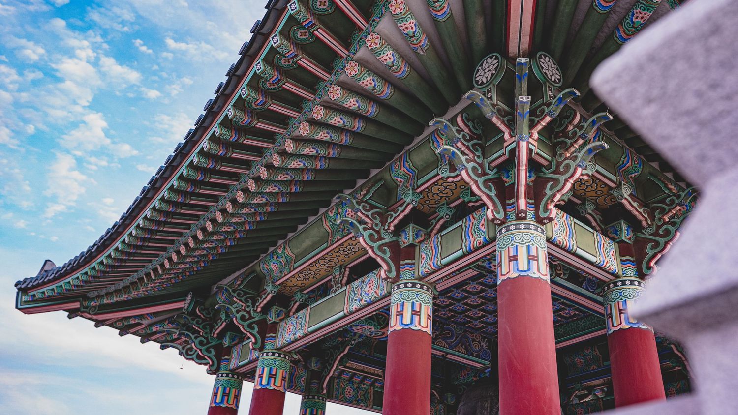 Ornate Korean traditional pavilion with colorful painted details, red columns, and curved tiled roof against a blue sky.
