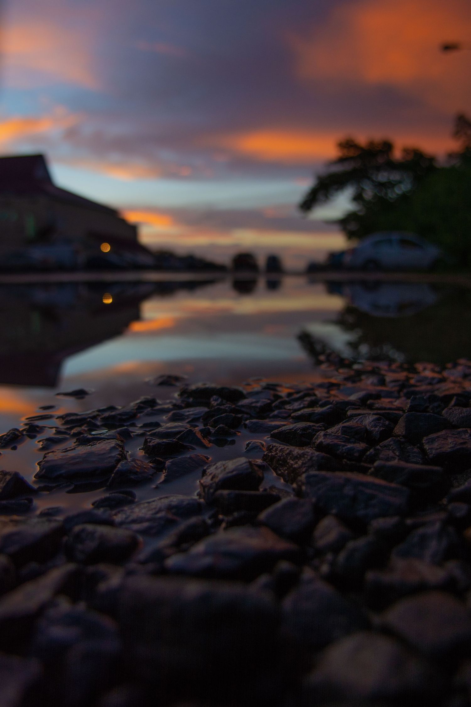 Dramatic sunset sky reflected in rocky shoreline with silhouetted objects in background.