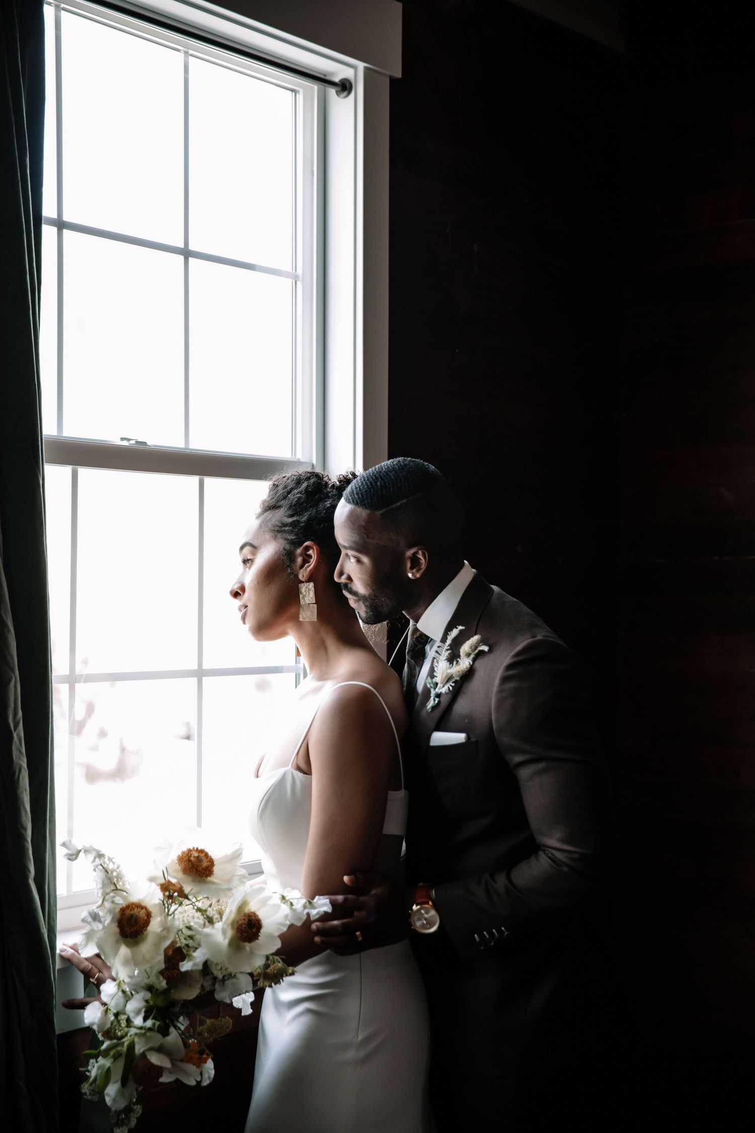 Silhouetted couple shares an intimate moment by large windows in formal attire.