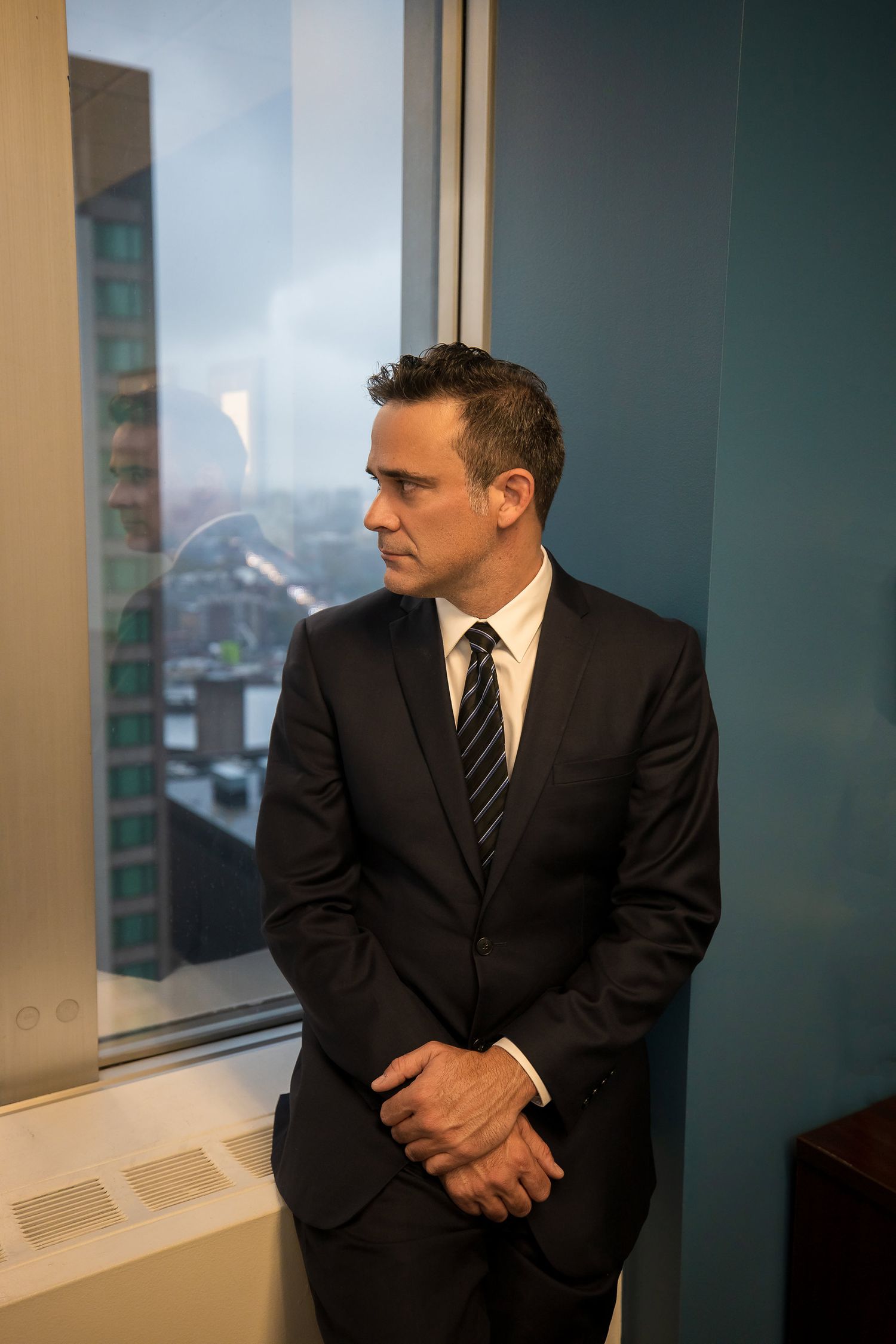 Professional in dark suit sits contemplatively by office window.