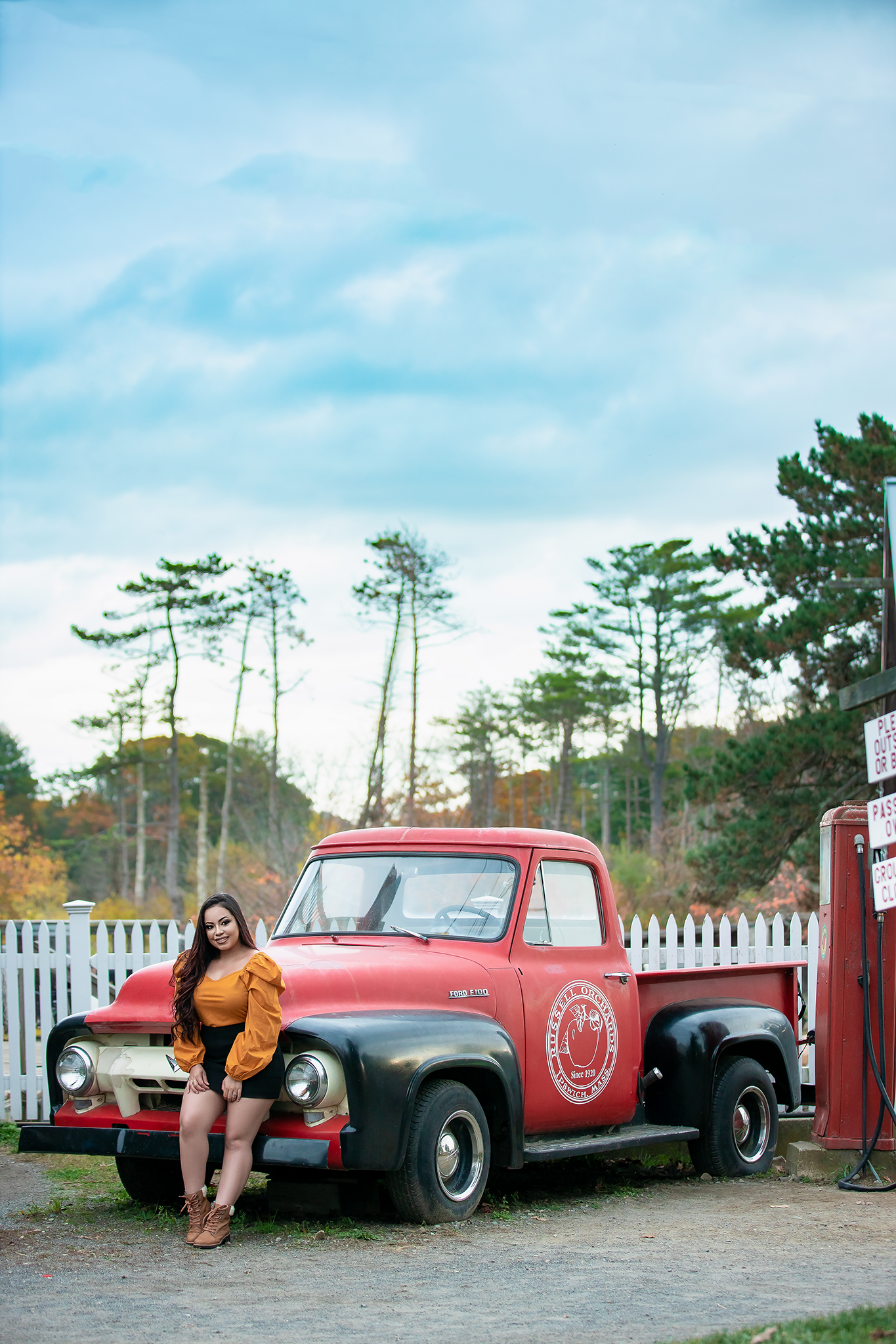 Vintage red pickup truck parked near pine trees and white fence.