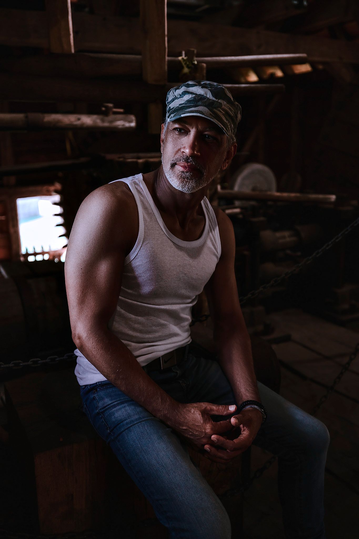 Person in white tank top and jeans sitting in dimly lit room.