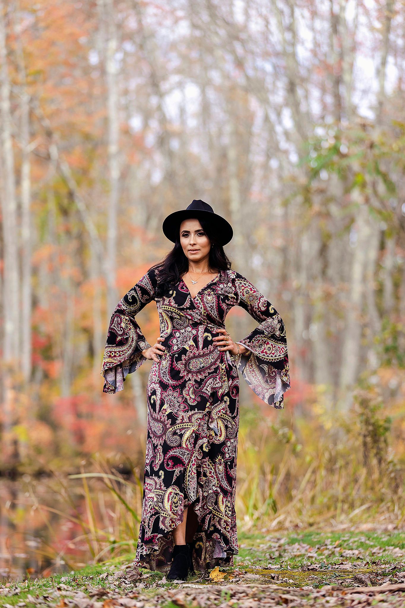 Outdoor fashion shoot in autumn woods featuring flowing patterned dress and black hat.