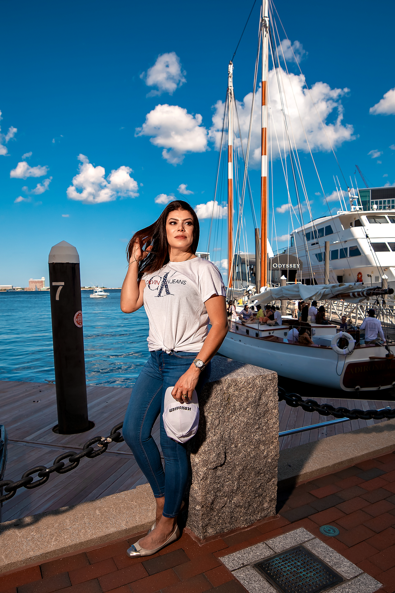 Person in casual attire posing by a marina dock with sailboats and blue sky in the background.