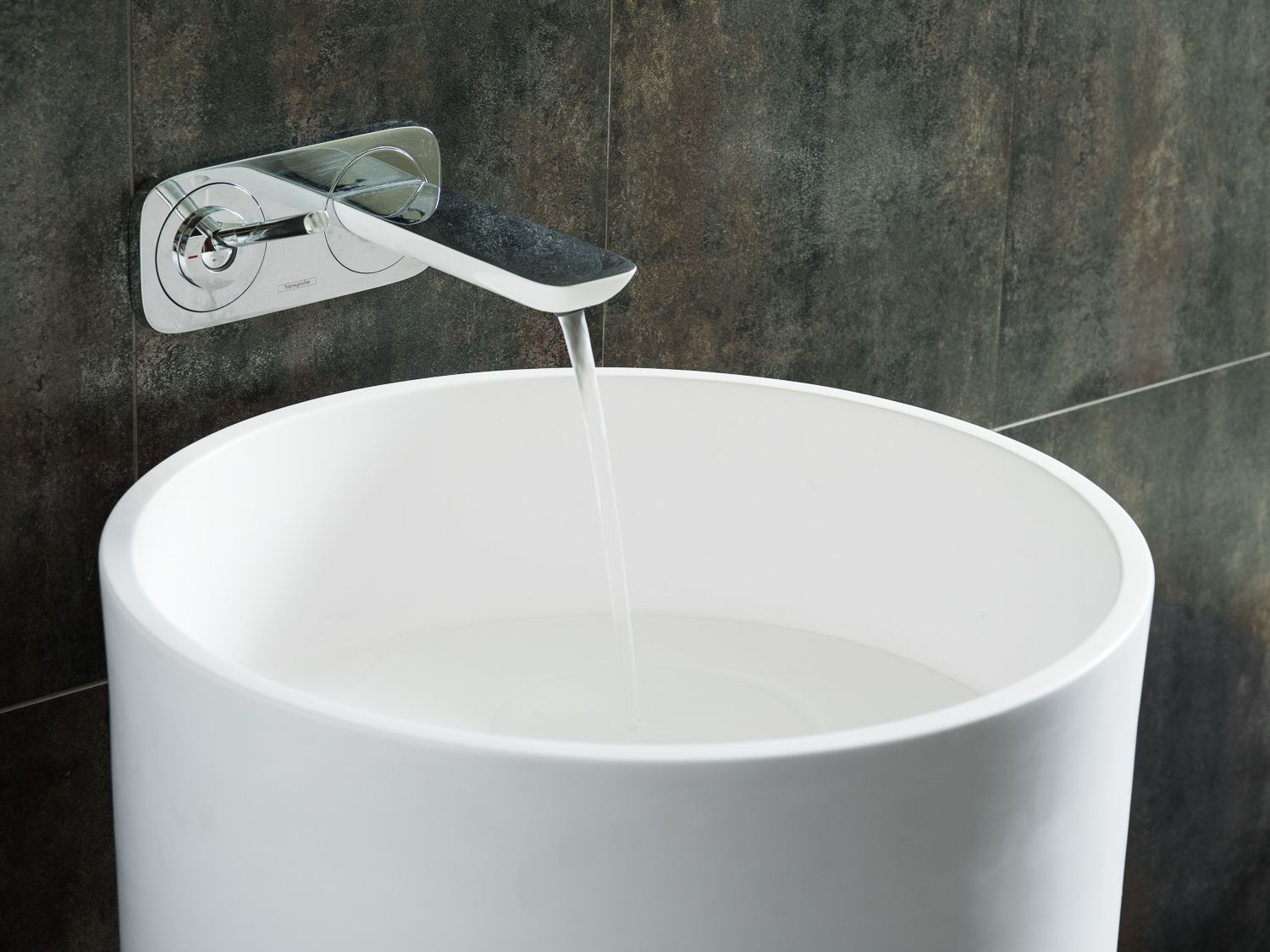 A modern white wall-mounted faucet pours water into a cylindrical vessel sink against a dark textured wall.