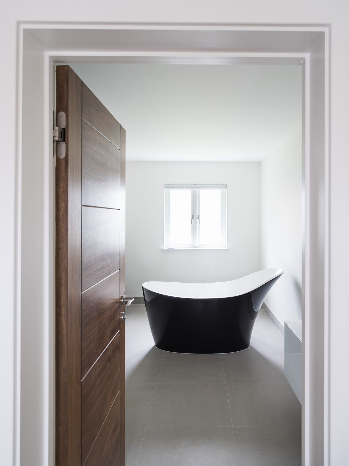 A modern bathroom features a sleek black freestanding tub against white walls with a wooden door and small window.