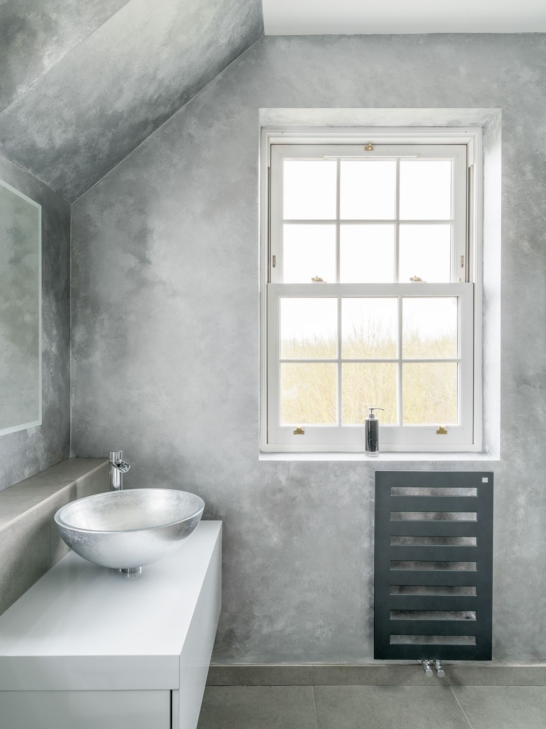 Modern bathroom interior with white vessel sink, grey concrete walls, and black radiator beneath bright window with field view.