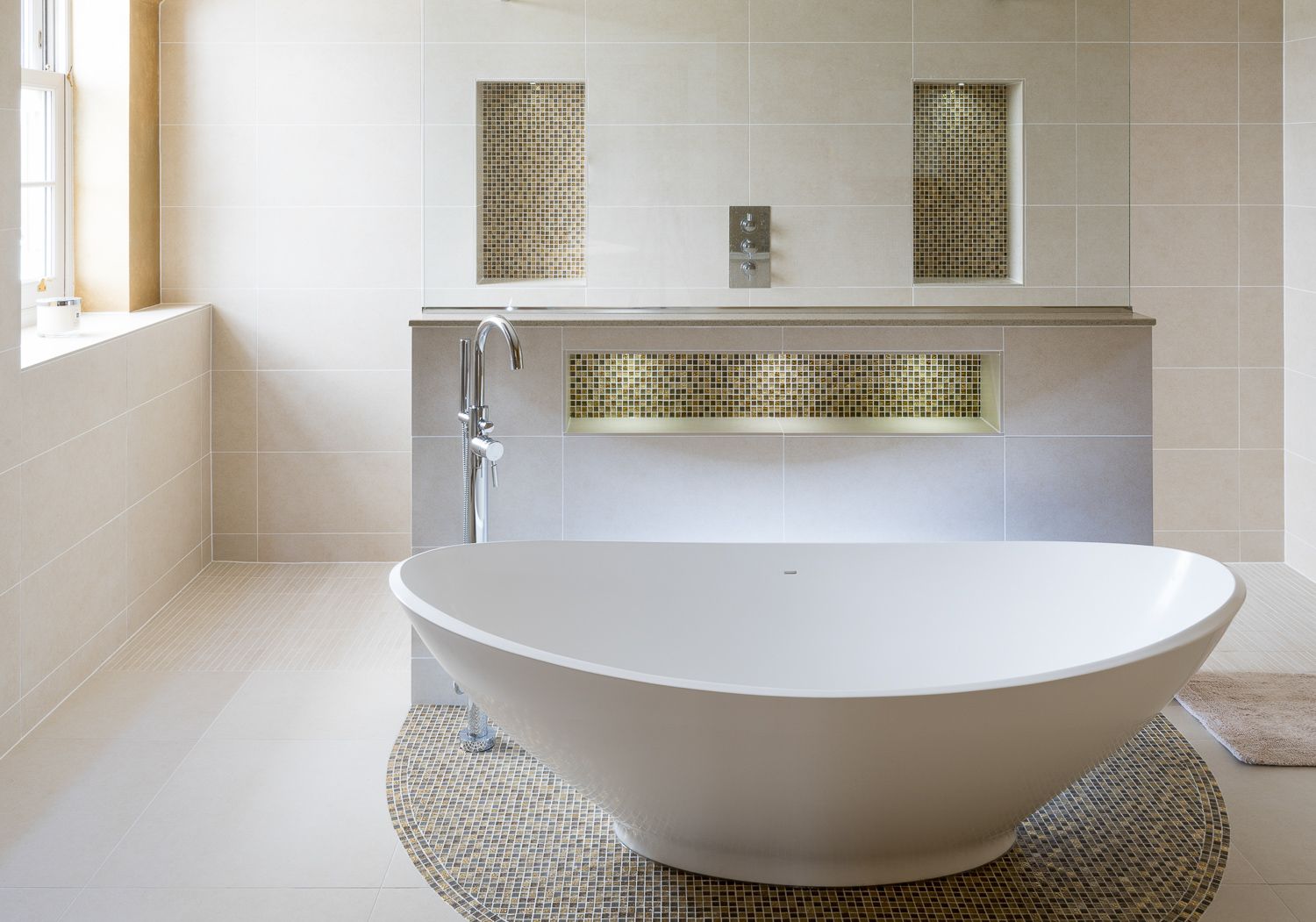 Sleek white oval freestanding bathtub on mosaic tile with gold accents and built-in wall niche in modern bathroom.