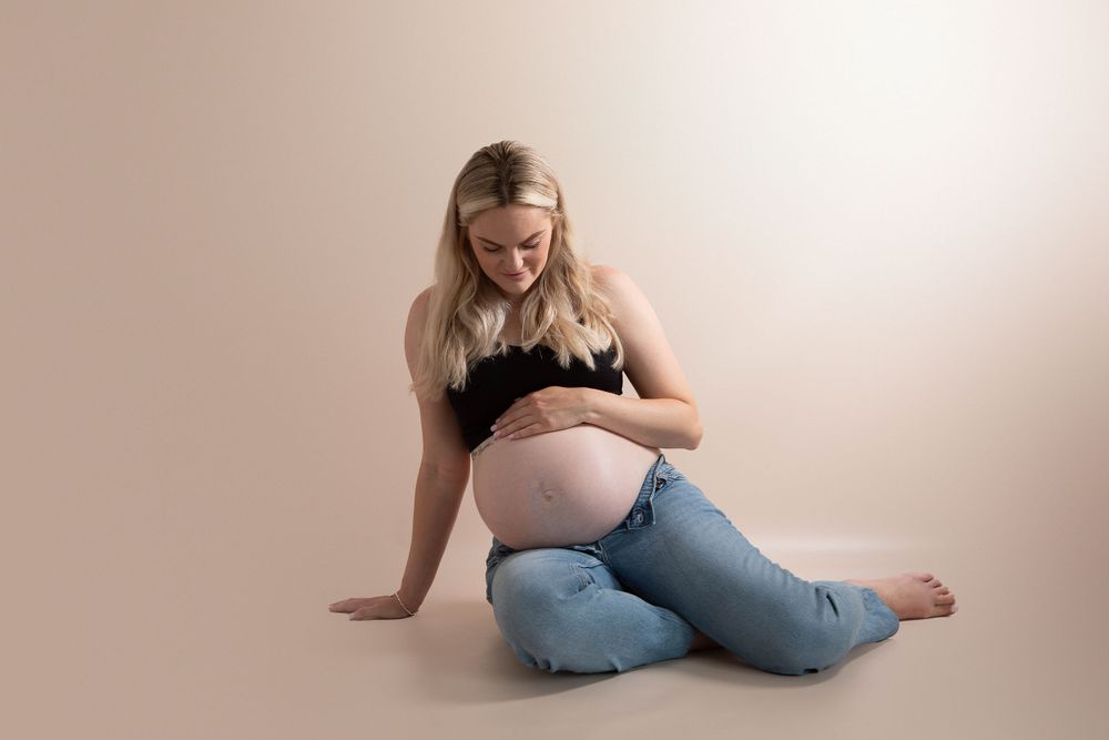 Pregnant woman in black crop top and jeans sitting on floor cradling baby bump in studio.