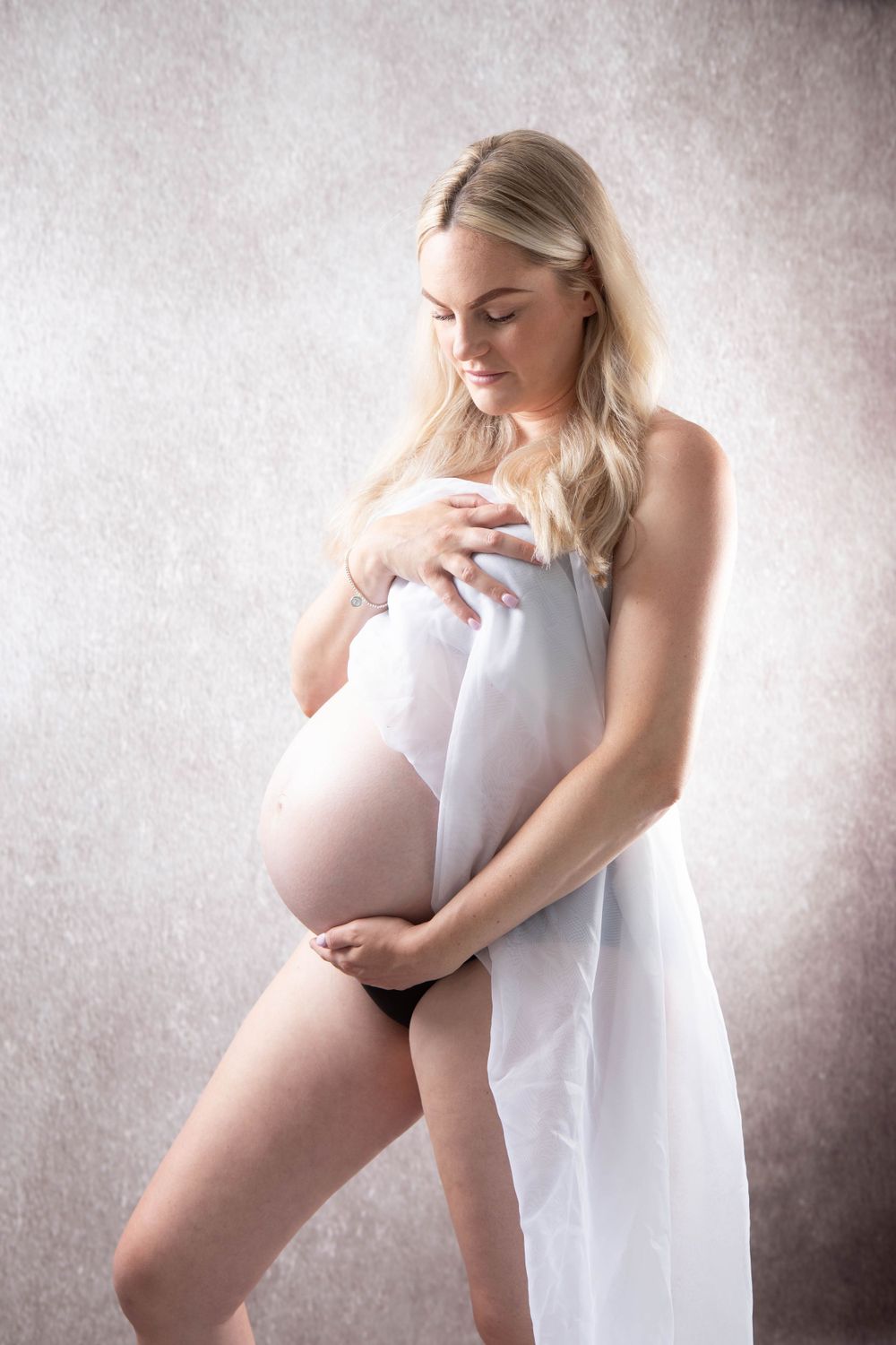 Blonde pregnant woman wrapped in white fabric holding baby bump in artistic maternity portrait.