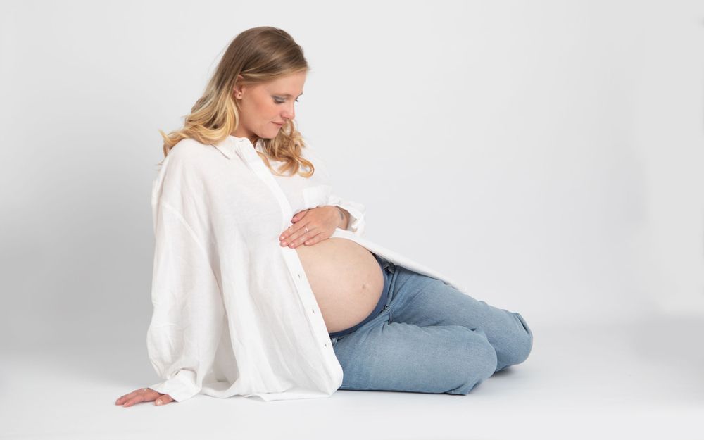 Pregnant woman in white shirt and jeans sitting on floor gently holding her baby bump.