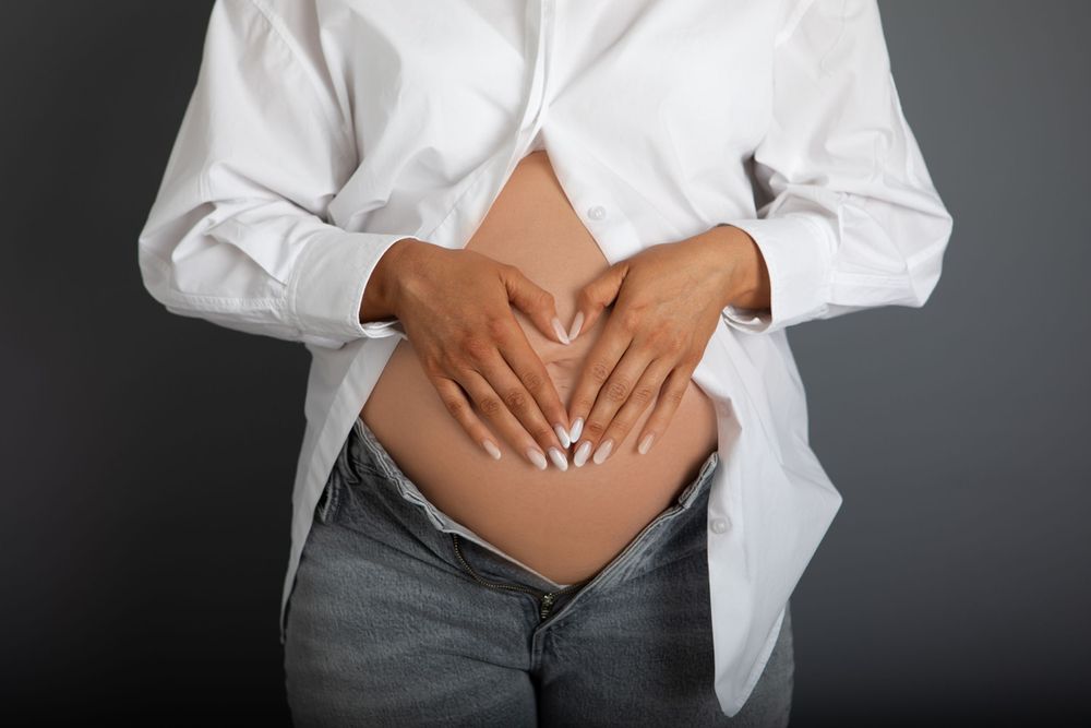 Close-up of pregnant woman in white shirt forming heart shape with hands on bare baby bump.