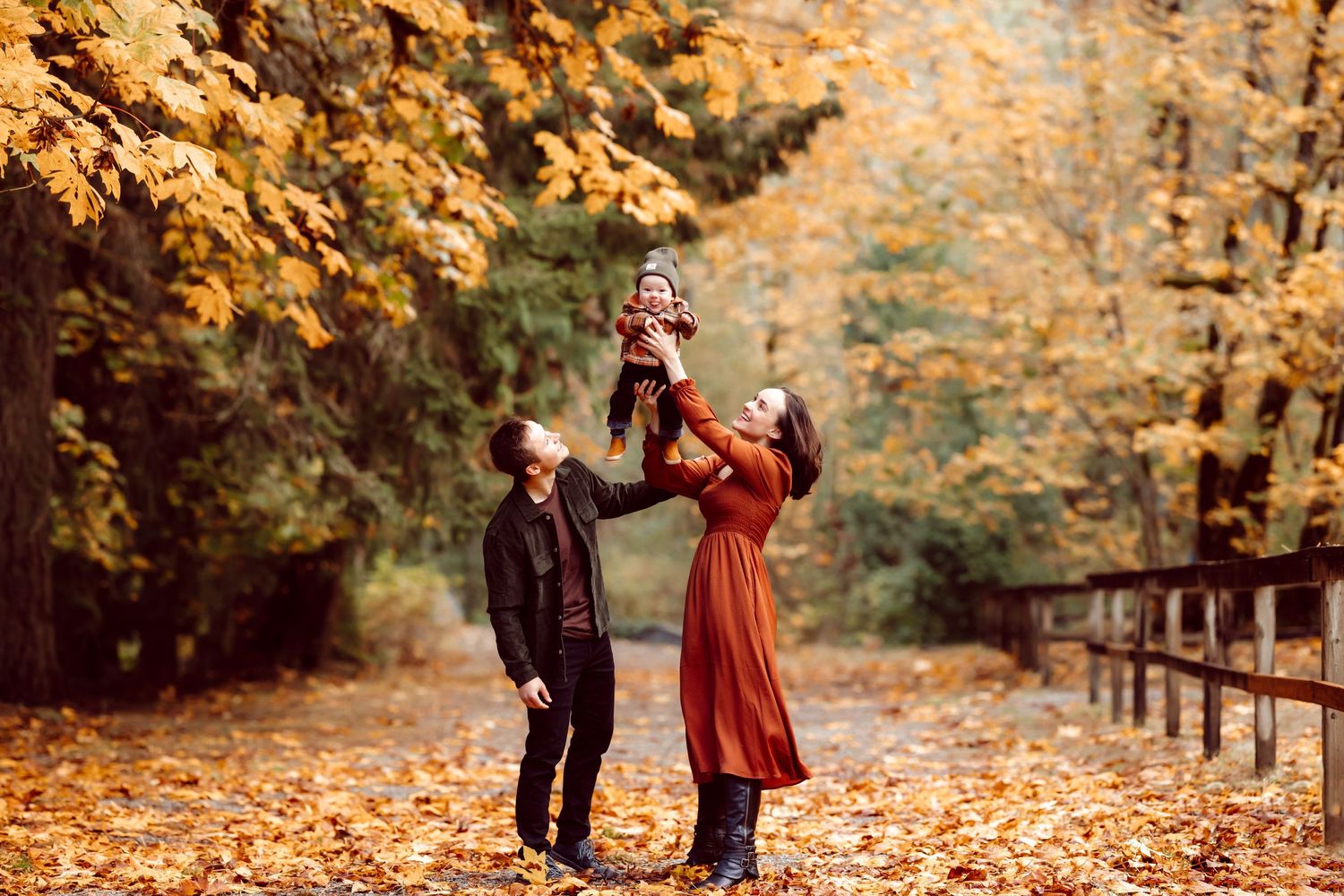 Family enjoying autumn scenery with golden leaves as parents lift child in playful moment.