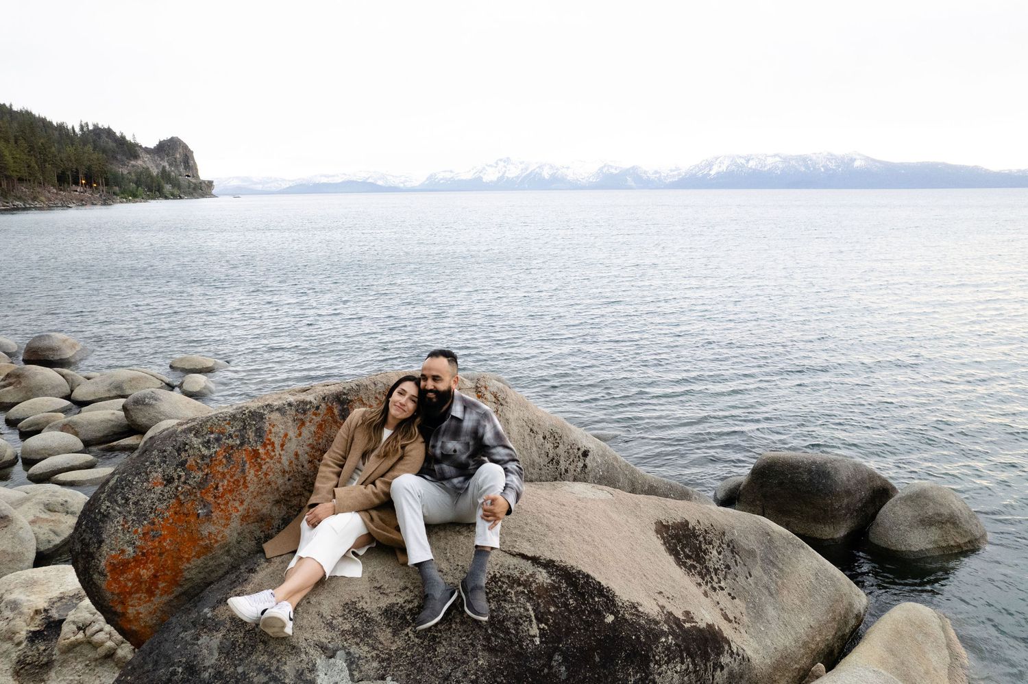 A couple sits together on large boulders along the shore of Lake Tahoe with snow-capped mountains in the distance.