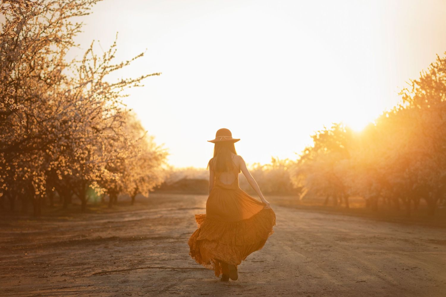 Silhouette of person in flowing dress dancing on dirt road at golden sunset.