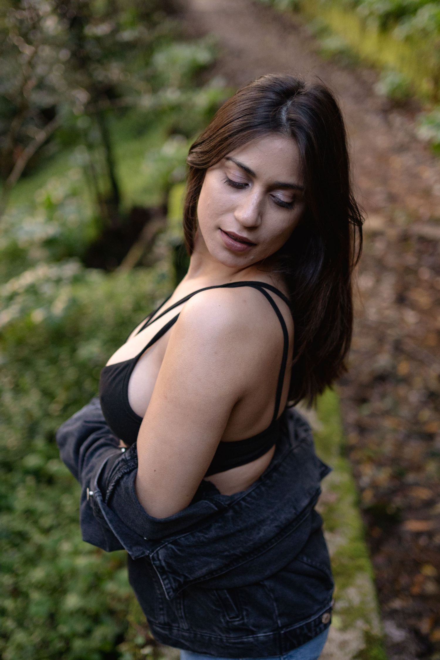 Portrait of woman in black top and denim jacket posing on forest trail.