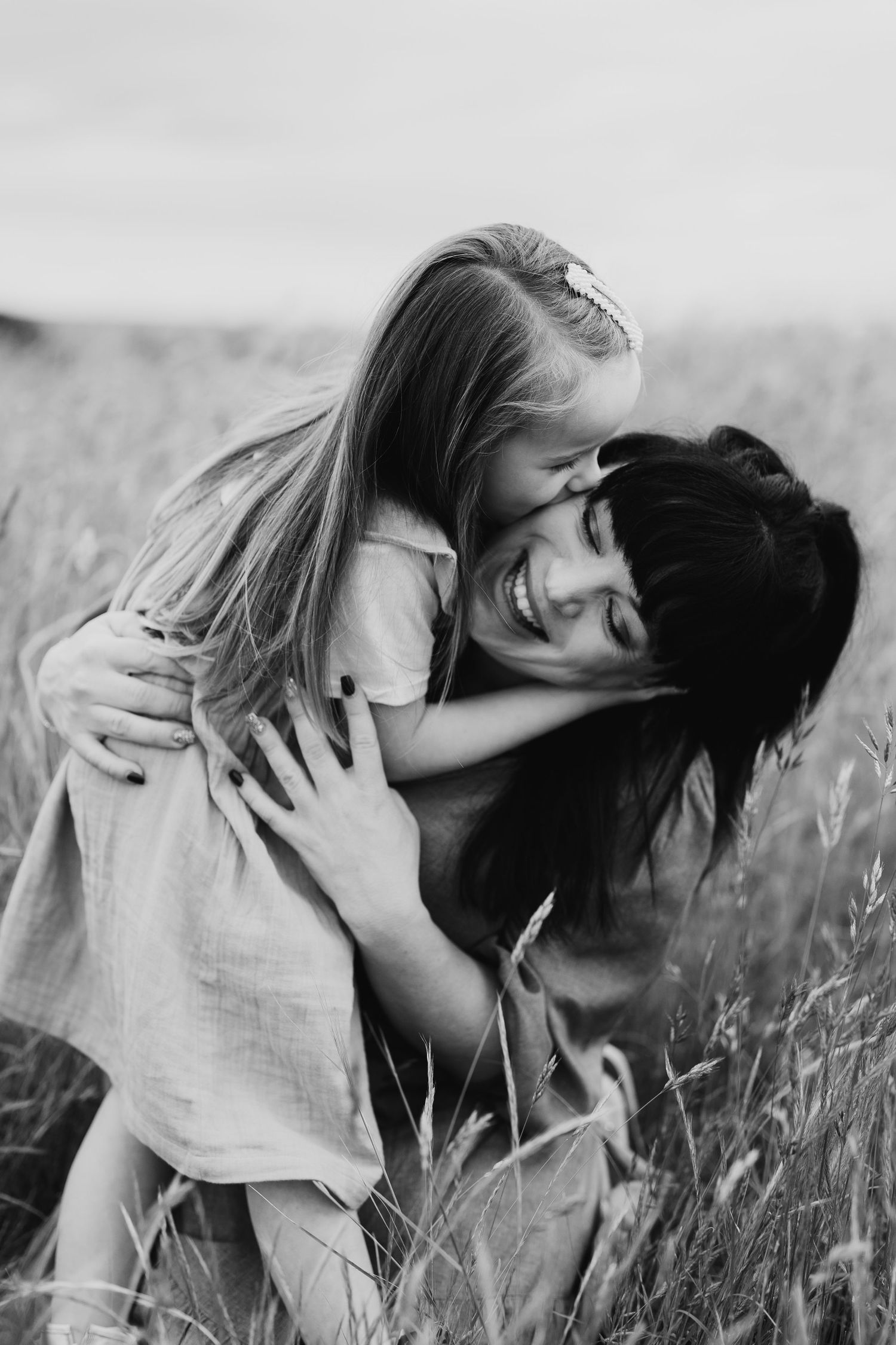Black and white photo of a smiling dark-haired mother hugging her blonde daughter in a field.