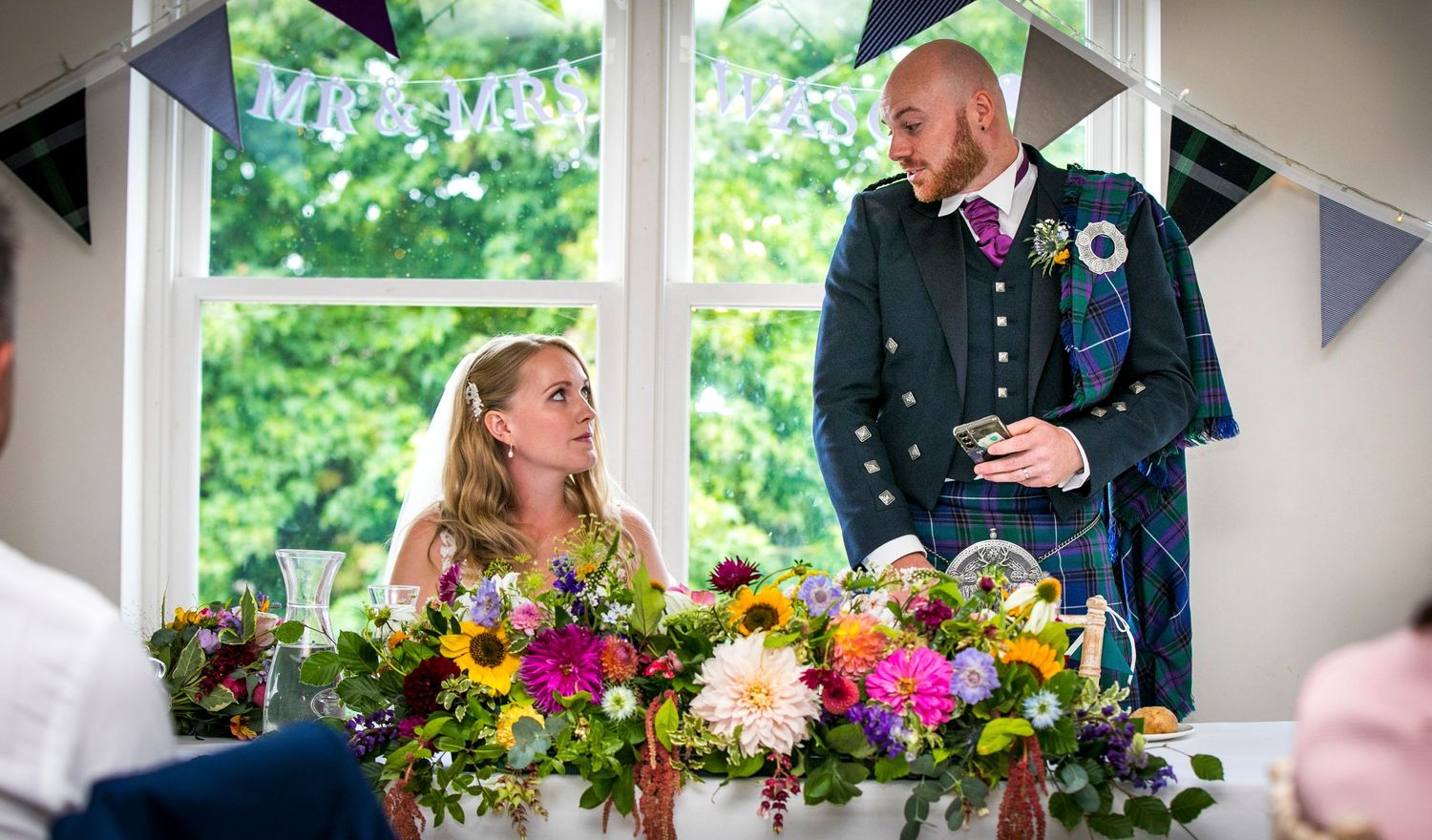 Wedding reception table decorated with vibrant yellow and pink flowers near large windows.