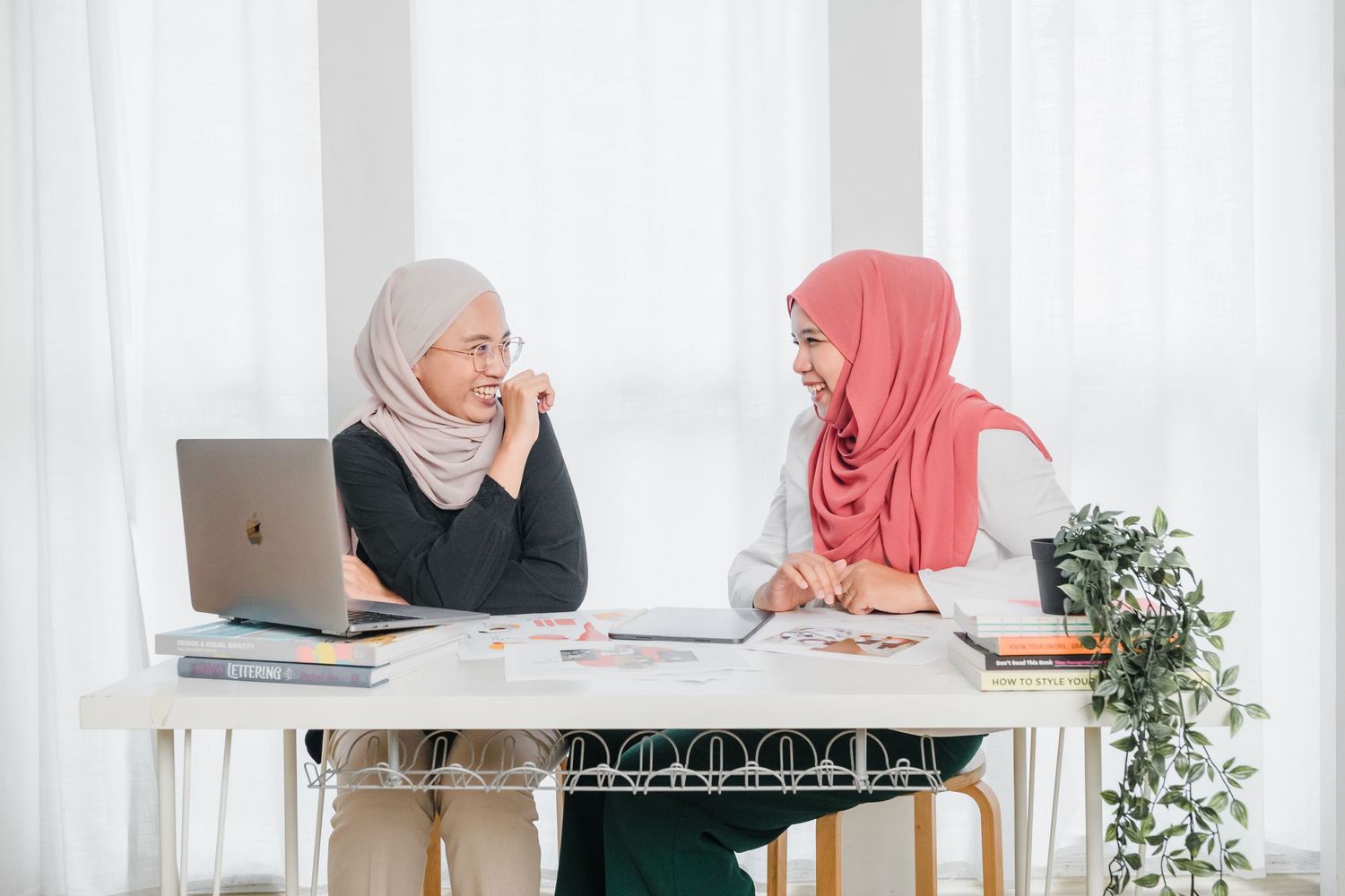 Two people in hijabs collaborate at desk with laptop and documents.