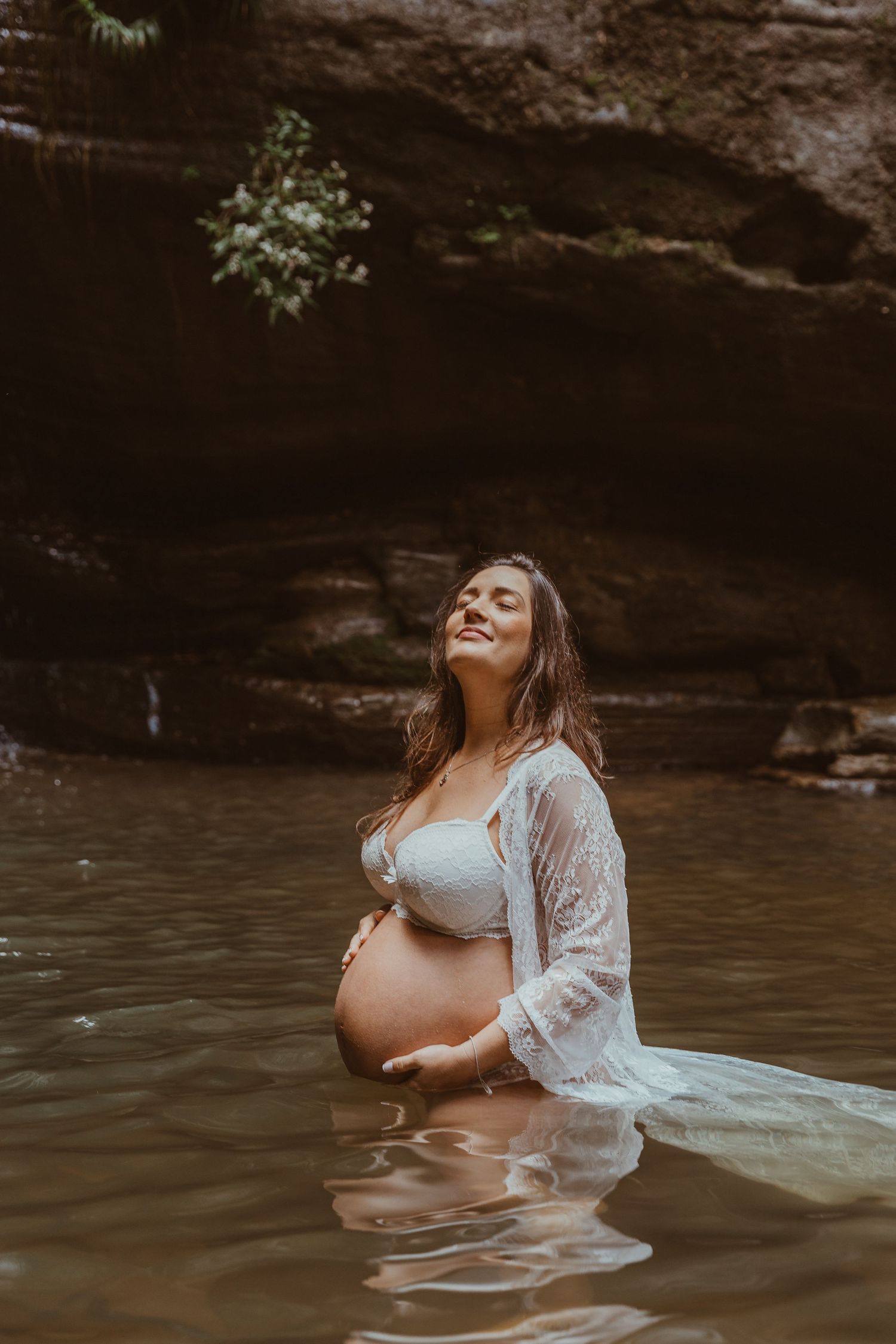 Ethereal maternity photo of pregnant woman in white dress standing in water near rock formation.