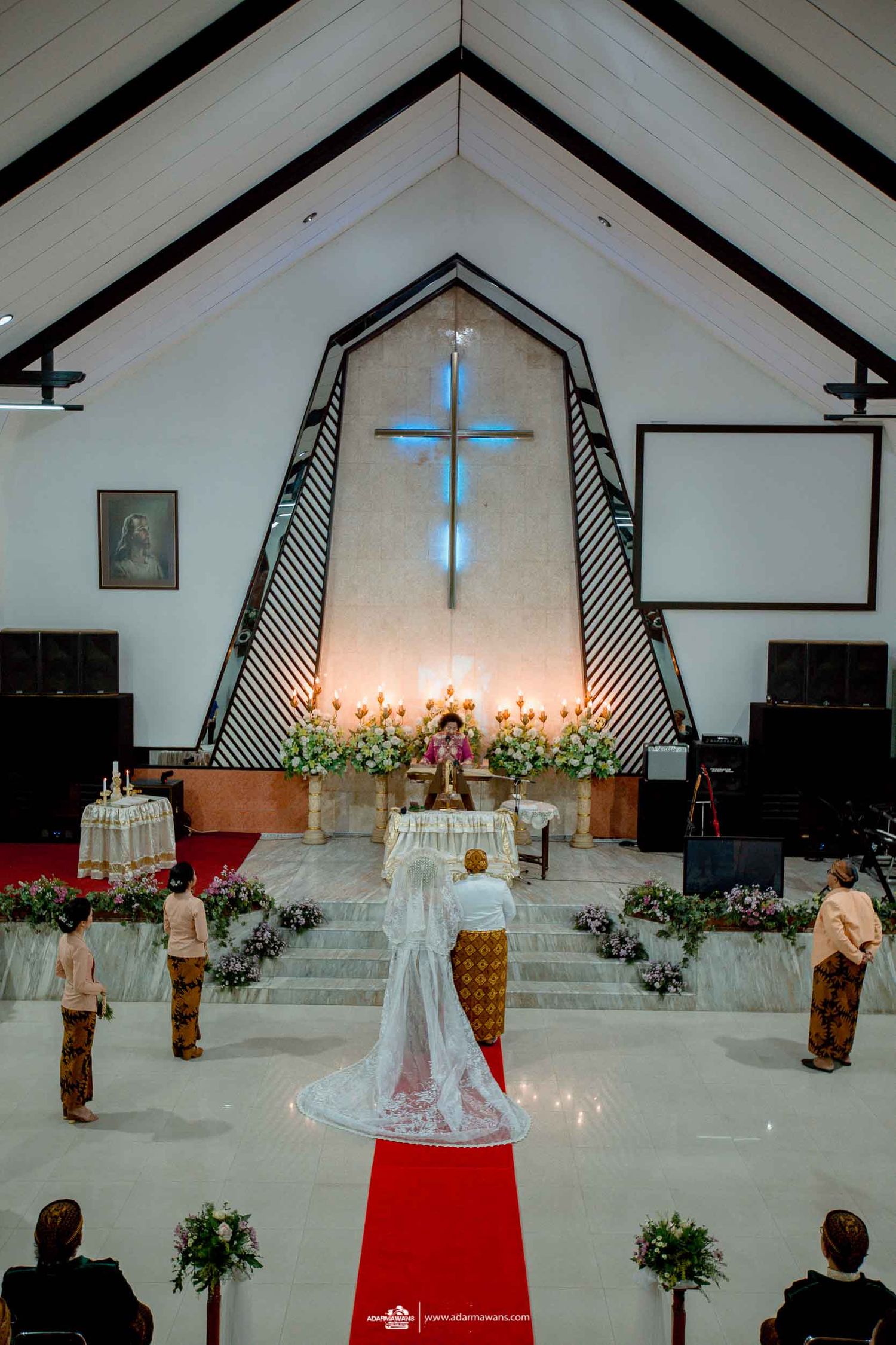 Church wedding ceremony setup with illuminated cross, candles and red carpet in a white chapel interior.