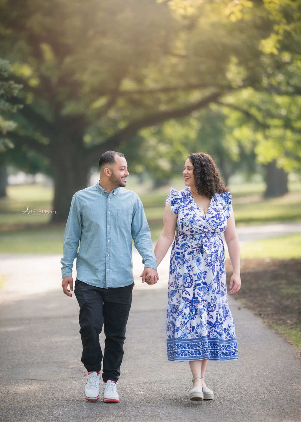 A couple strolls together on a sunlit park path, wearing coordinating blue outfits while holding hands.
