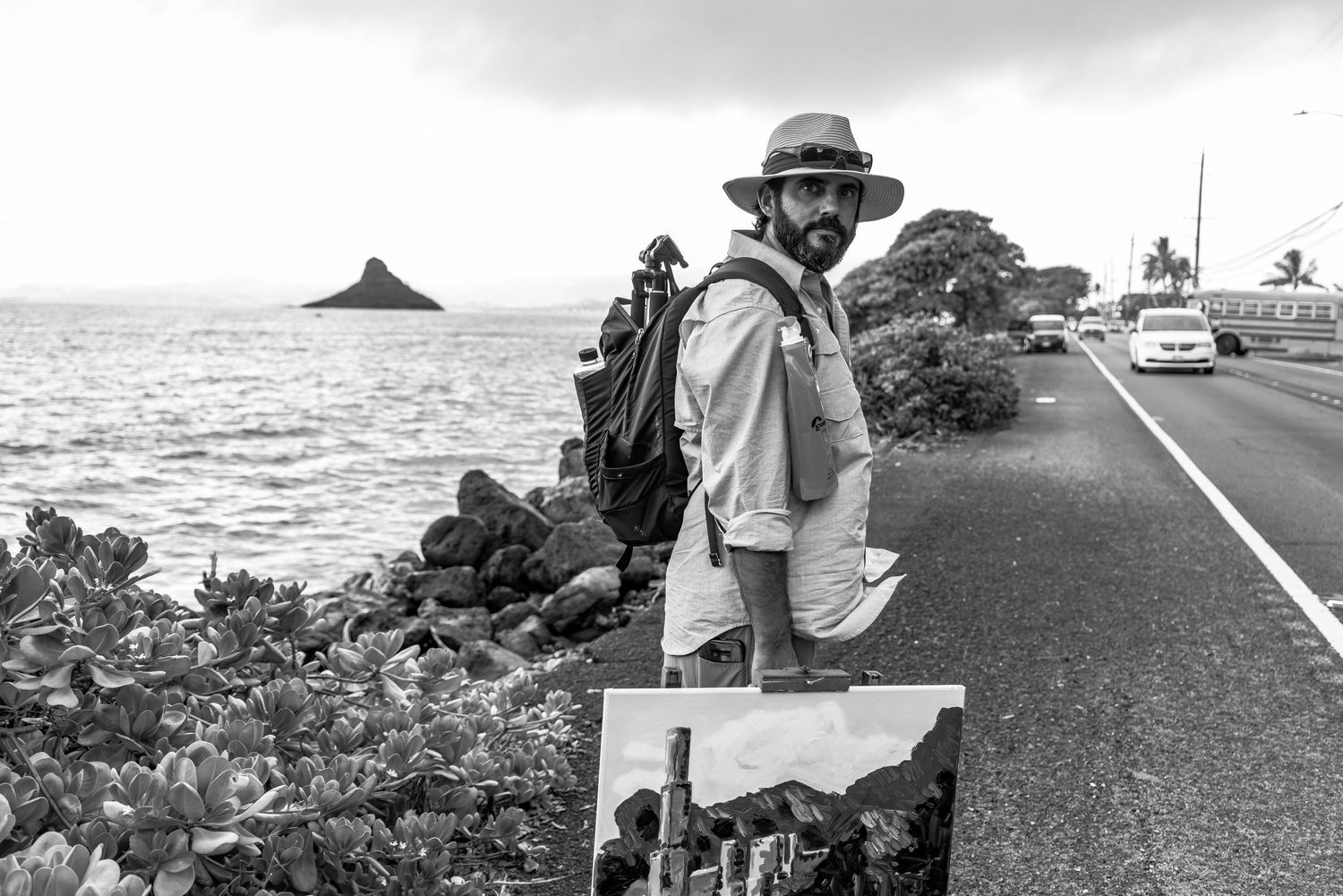Black and white photo of photographer holding artwork on coastal highway with ocean view.