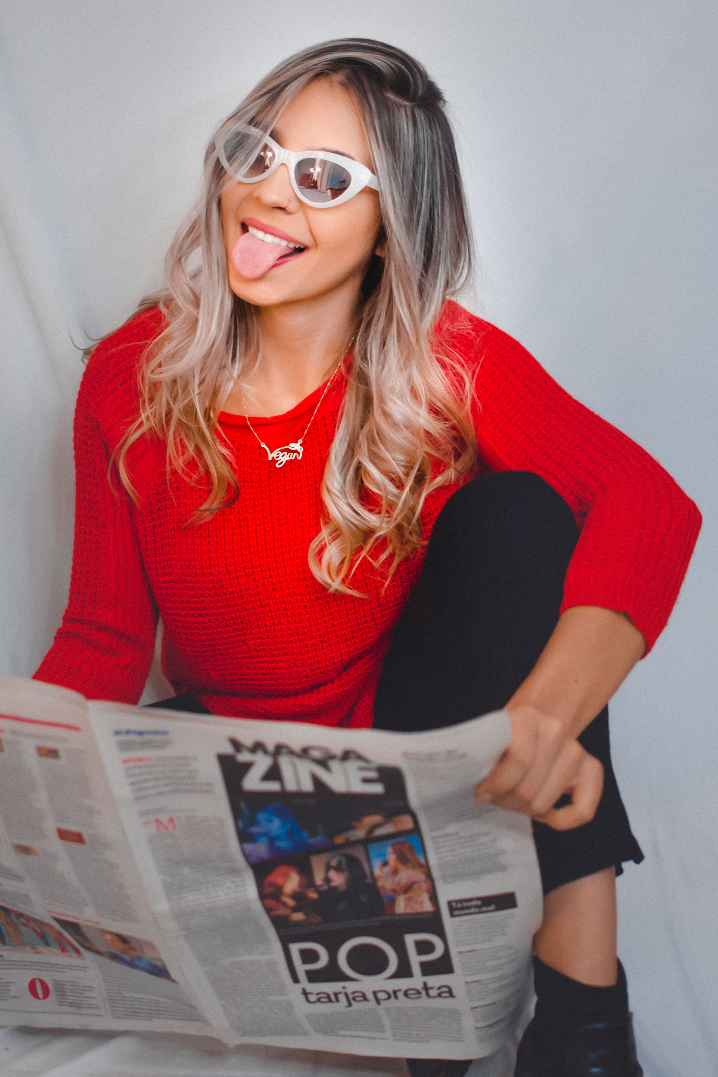 Playful portrait in red sweater reading newspaper while wearing retro sunglasses.