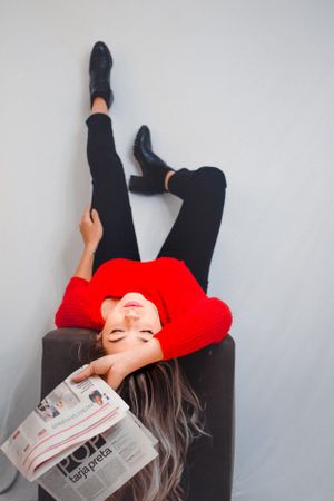 Person in red sweater lies on floor while reading newspaper against white background.
