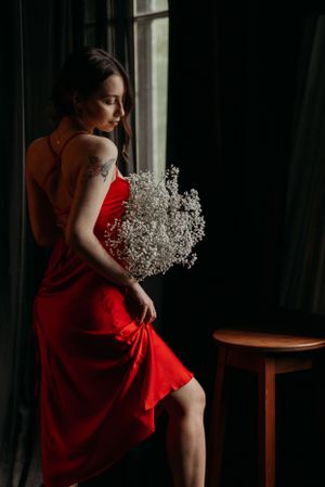 Dramatic portrait series in red dress with baby's breath flowers near window.