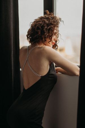 Person with curly hair in black top leaning against window in moody lighting.