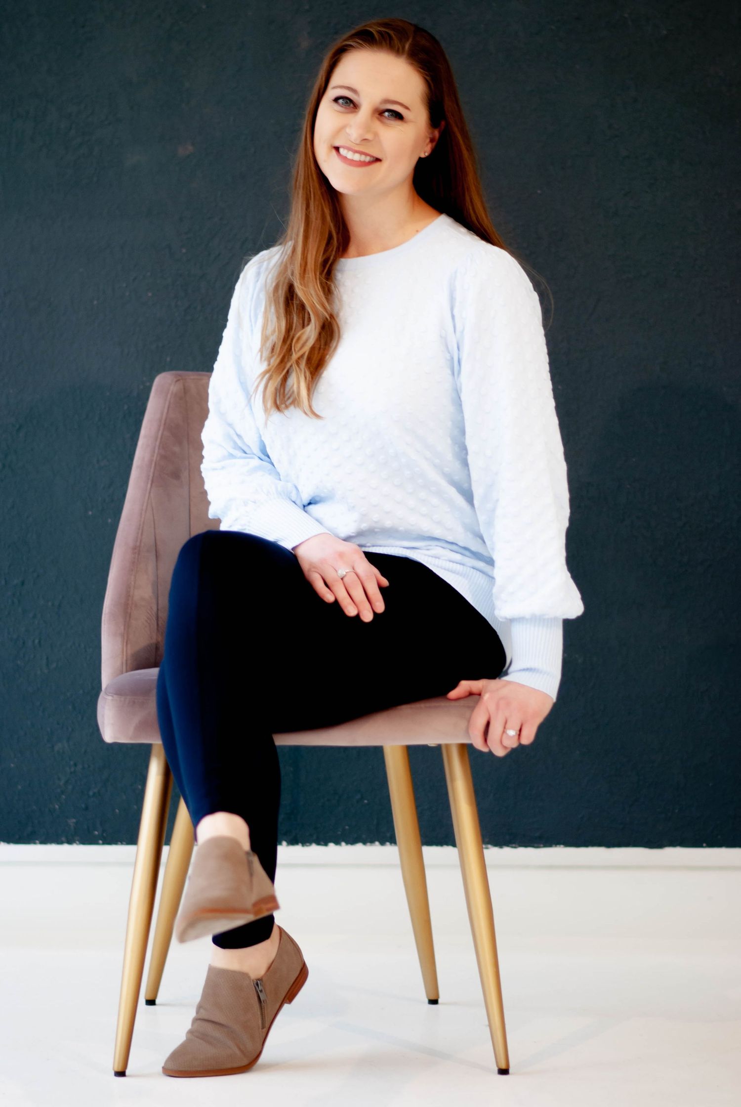 Smiling woman in light blue quilted sweater and navy leggings seated on a pink chair.