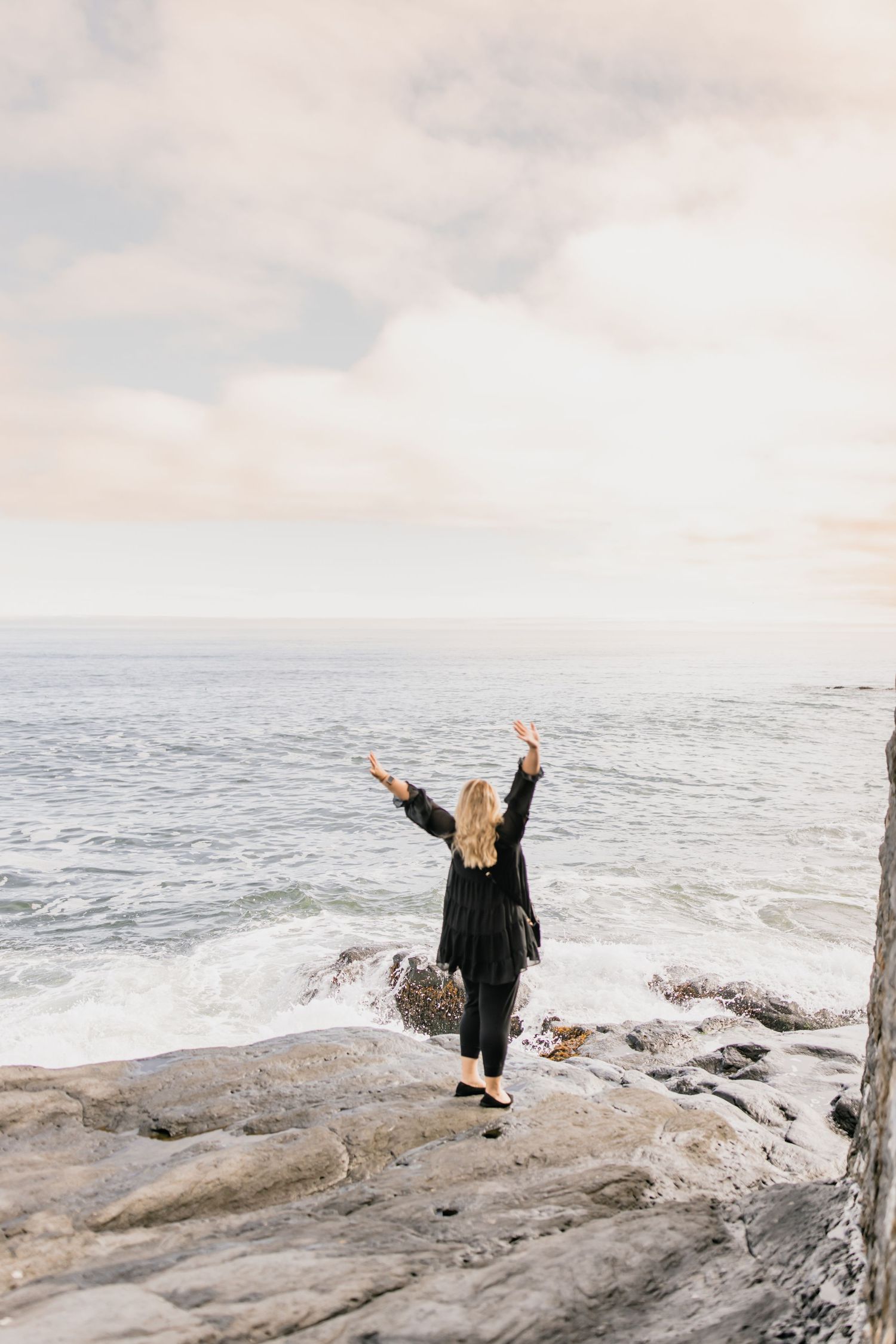 Blonde woman in black outfit standing on rocky cliff with arms raised toward ocean.
