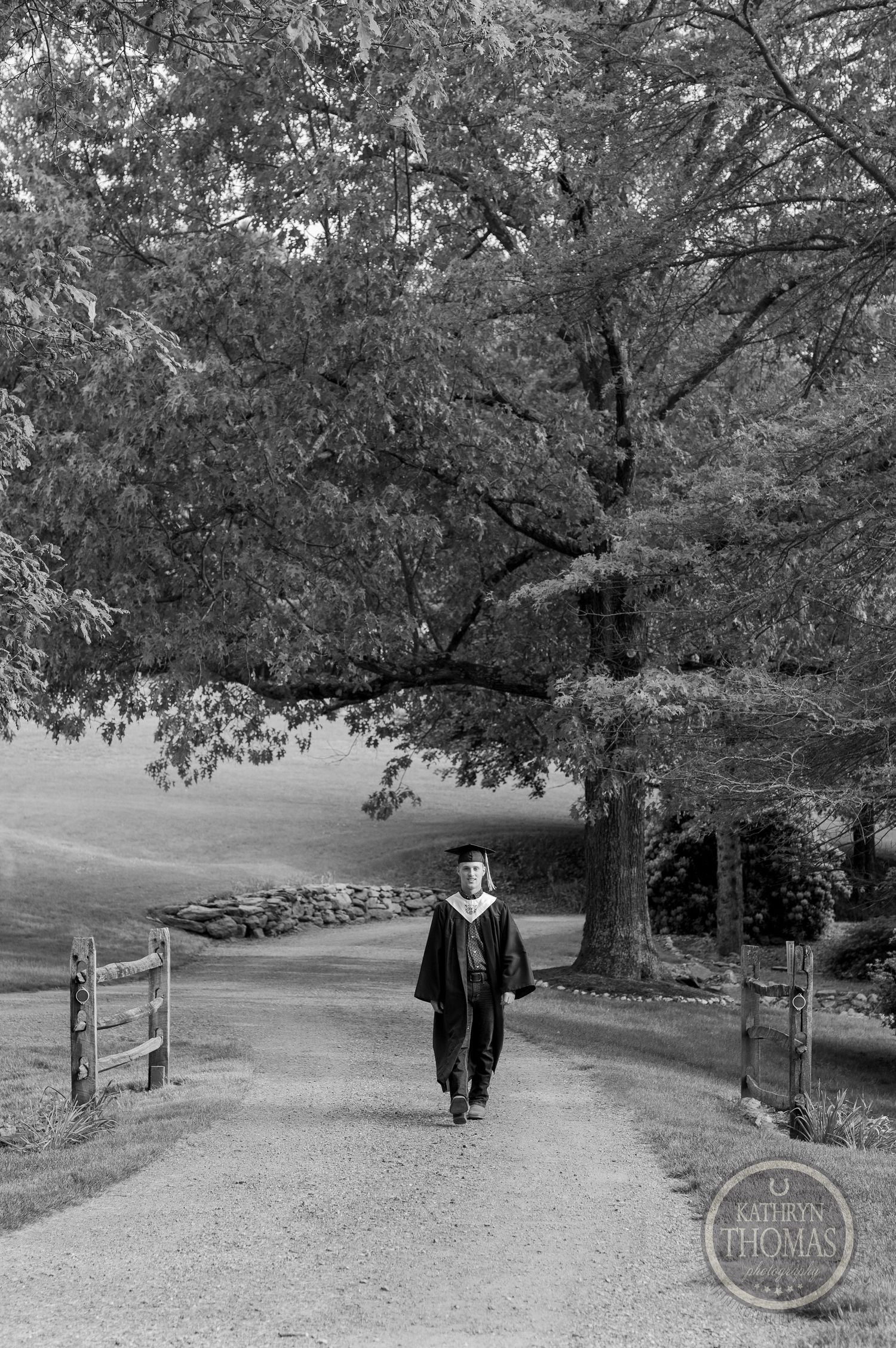 Black and white photo of a graduate walking down a tree-lined path in academic regalia.