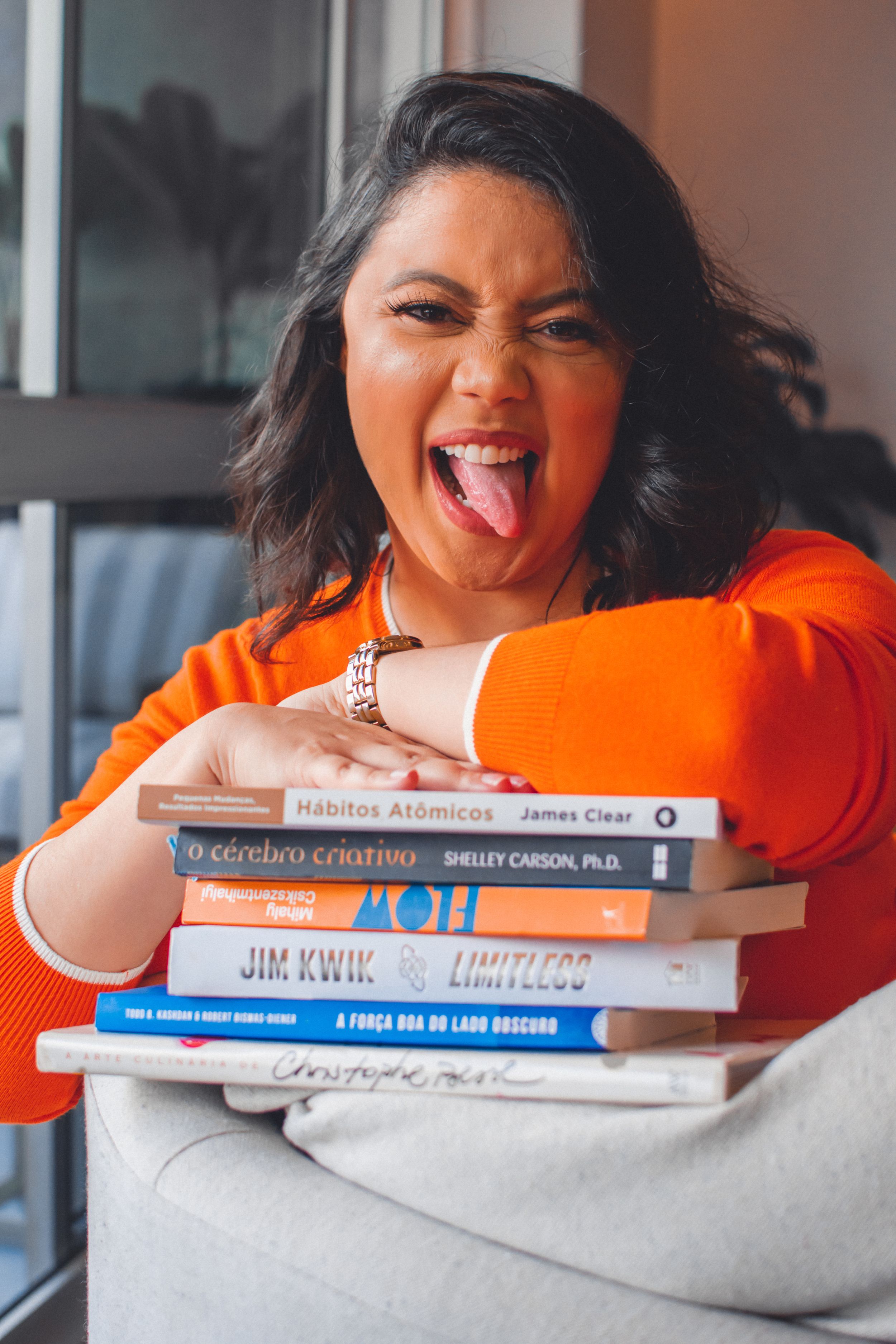 Cheerful poses with a stack of self-help and motivational books in an orange sweater.