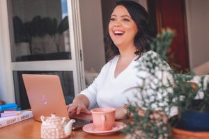 Person working on laptop with tea and plants on desk, smiling brightly.