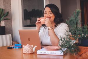 Person smiling while enjoying tea at desk with laptop and books.